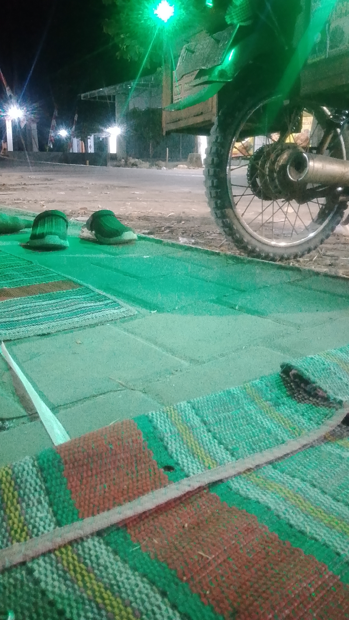 At night in Karanglangu, Indonesia, a scene unfolds on a paved area adjacent to a road. In the foreground, a woven mat with stripes of red, green, and yellow lies on the pavement. Two green shoes are placed side-by-side on a green mat nearby. Behind the mats, a motorcycle is parked. The rear wheel of the motorcycle is prominently visible, with its spokes and tire tread. The motorcycle's exhaust pipe is also visible, along with a portion of its body. A bright green light emanates from above, casting a glow on the scene. In the background, there are some buildings and structures, and lights from street lamps are visible, suggesting an urban or semi-urban environment. The overall impression is one of a quiet night setting, possibly in front of a residence or a roadside establishment.