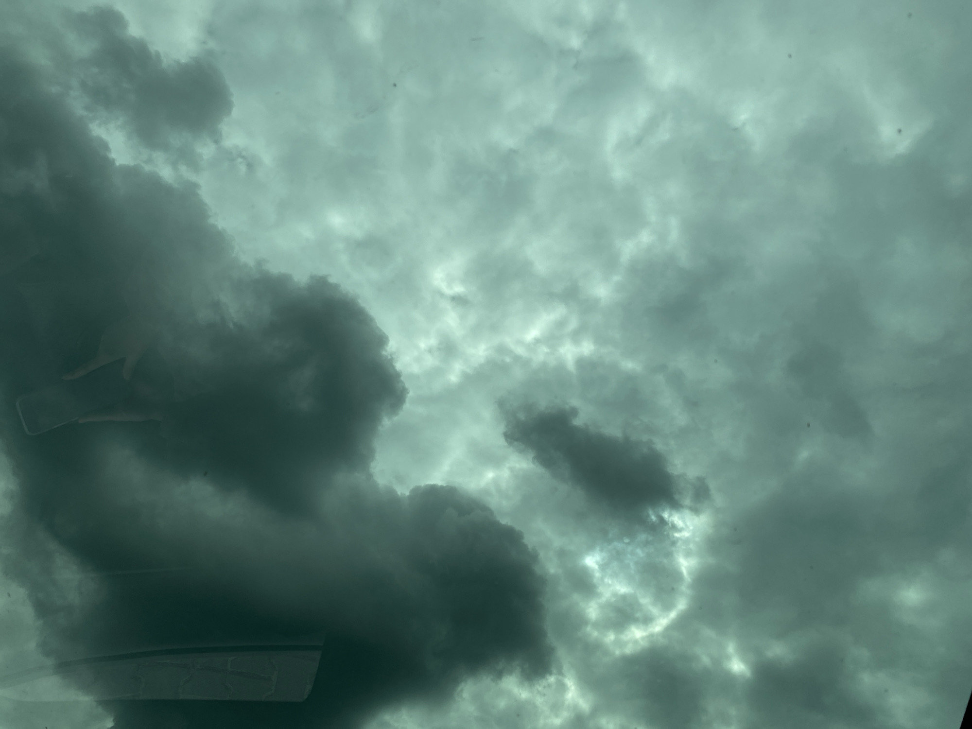 The image shows a view of a cloudy sky, seemingly taken from inside a vehicle. The clouds are predominantly dark gray and white, suggesting stormy weather. There are no discernible people or specific objects in the scene apart from the clouds. The location is not identifiable within the city of Unknown, Unknown.