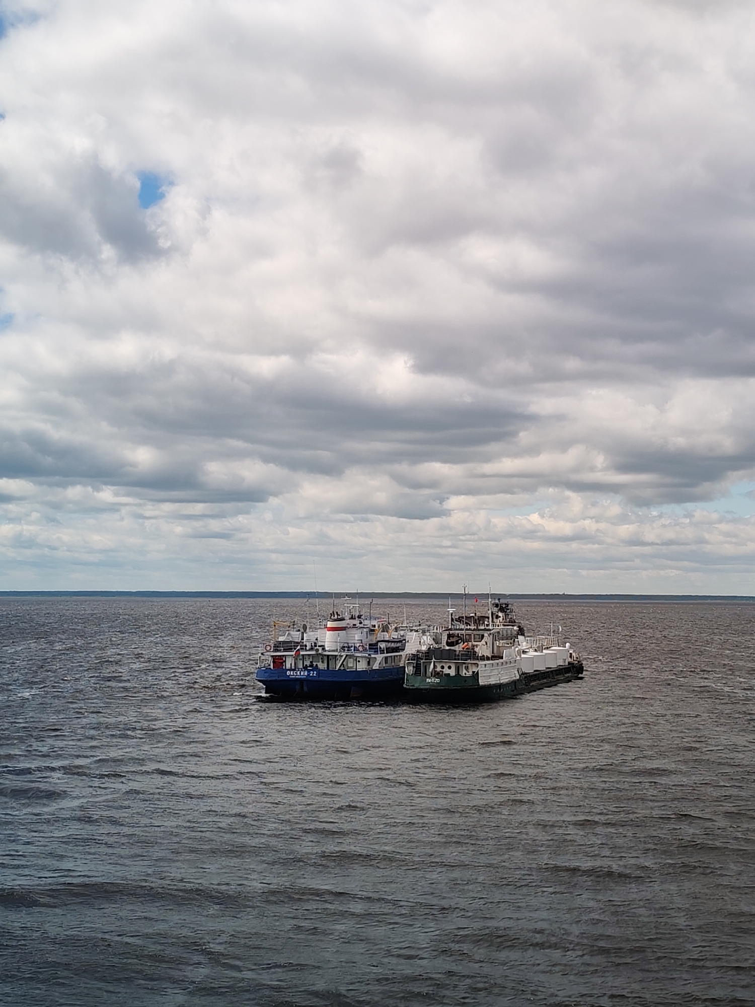 The image shows two ships or barges connected in the middle of a large body of water under a cloudy sky. The water appears dark, possibly due to lighting conditions or water color.
The ship on the left has a blue and white hull, and the name "OKCWM-22" is visible on its side. The ship on the right has a green and white hull. The name "TRIZO" is visible on its side. They are docked together.
The sky is filled with many layers of gray and white clouds, and there's a small patch of blue sky is visible through the clouds. The horizon is visible in the background between the water and the sky.