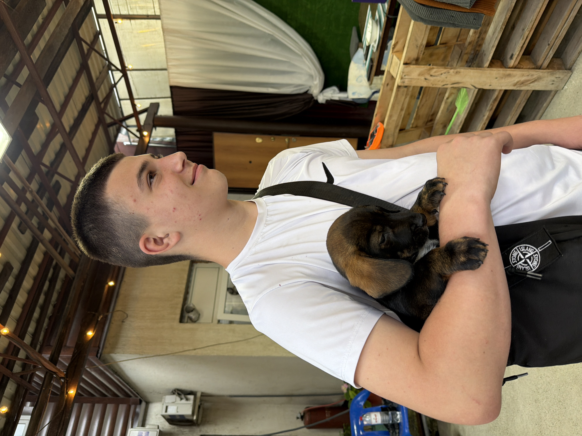 A young person with short brown hair and light skin, wearing a white t-shirt and a black crossbody bag, is holding a small brown and black puppy. The puppy is nestled in the crook of the person's arm, with its head resting on the person's arm. The background appears to be an outdoor covered area, possibly a patio or a market stall, with wooden structures, a corrugated roof with lights, and curtains. There are also some chairs and tables visible. The scene has a casual, relaxed atmosphere. The image is taken from a medium distance, focusing on the person and the puppy. The lighting suggests it is daytime. The location is stated to be Tuapse, Russia.
