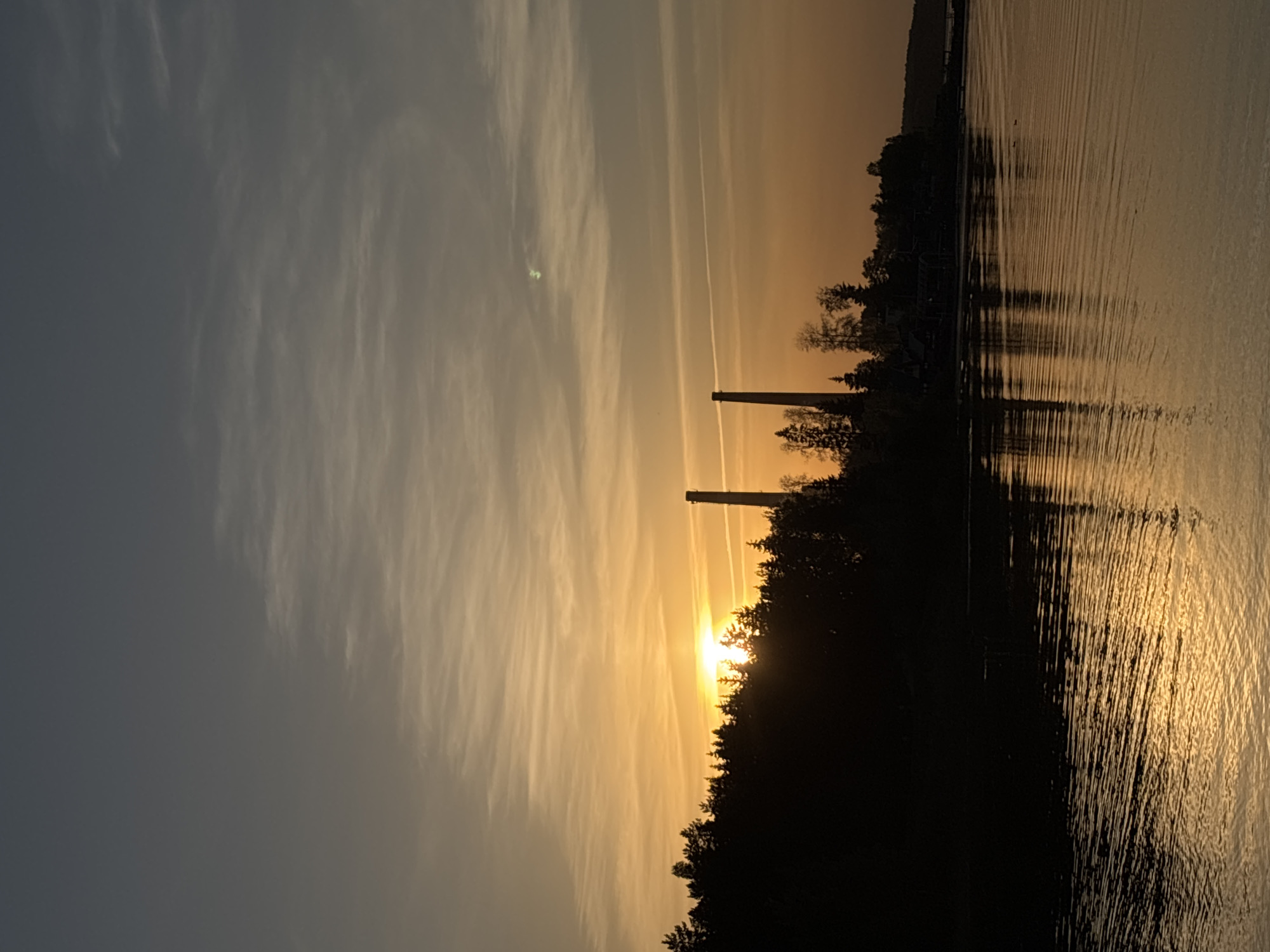 The image captures a sunset scene on the Kama River in Chaikovsky, Russia. The sun is setting behind a silhouette of trees and two industrial chimneys, casting a golden glow on the water's surface. The sky is a mix of gray and orange hues, with wispy clouds and contrails visible. The river's surface reflects the sunlight, creating shimmering patterns.