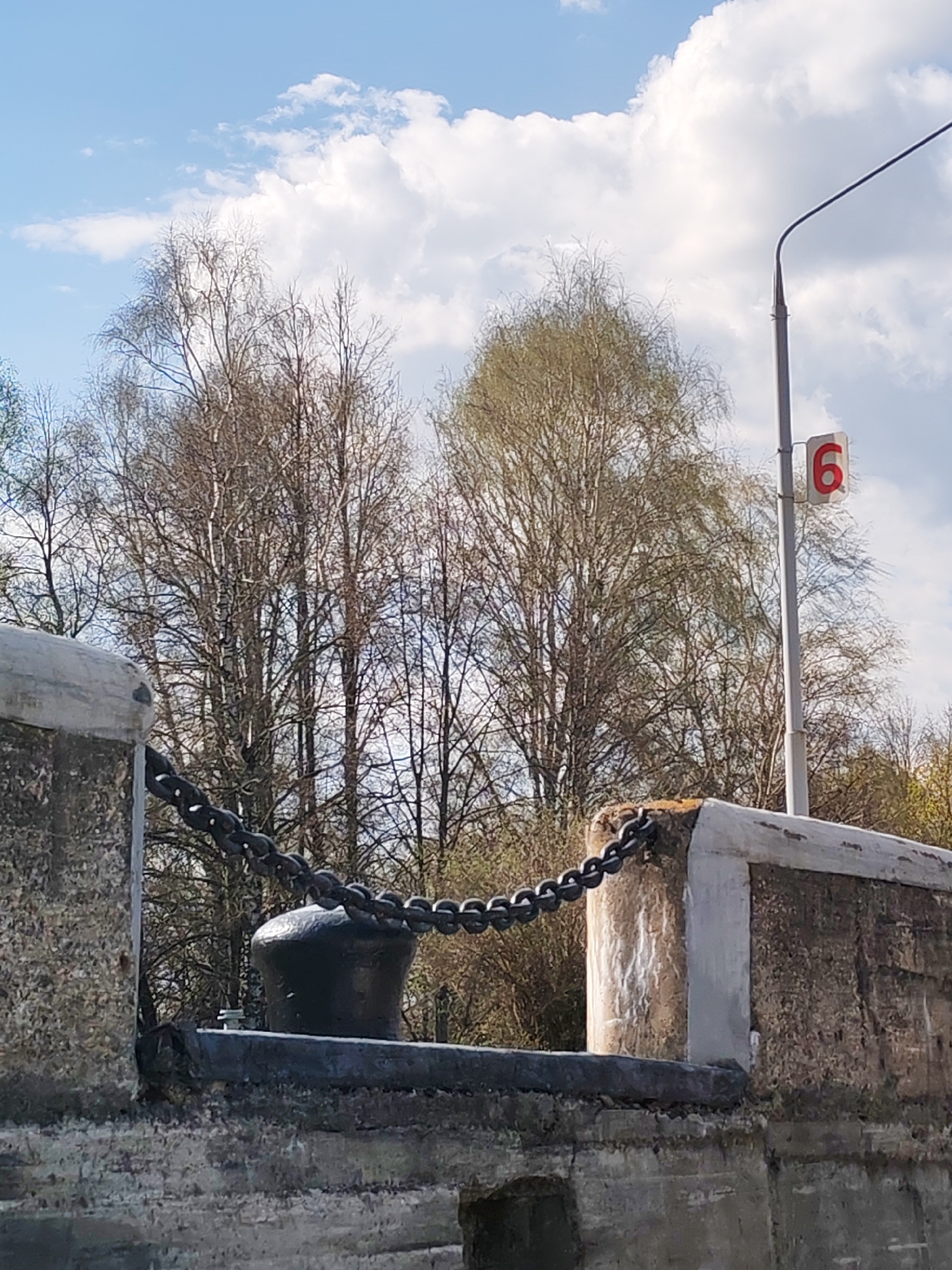 A street-level, medium shot shows a bridge lock in the city of Yakhroma, Russia. A black chain is strung between two concrete bridge barriers. A black metal mooring bollard sits on a black support beam, which is part of the concrete barrier. A white pole with a red sign "6" is standing behind the right bridge barrier. Several bare trees are standing behind the bridge lock. Light, white clouds are in the blue sky.