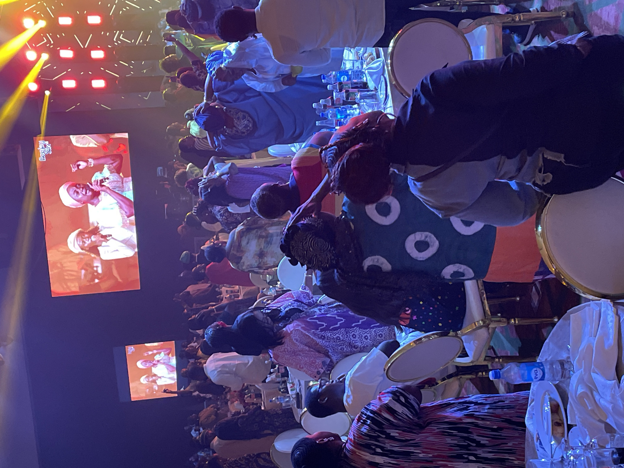An indoor gathering of people is captured in Itirin, Nigeria. Individuals are seated at tables, with chairs made of white material and gold trim. Some tables hold water bottles. Two large screens display a performance, showing two singers, one with a microphone. Overhead, stage lighting is visible.