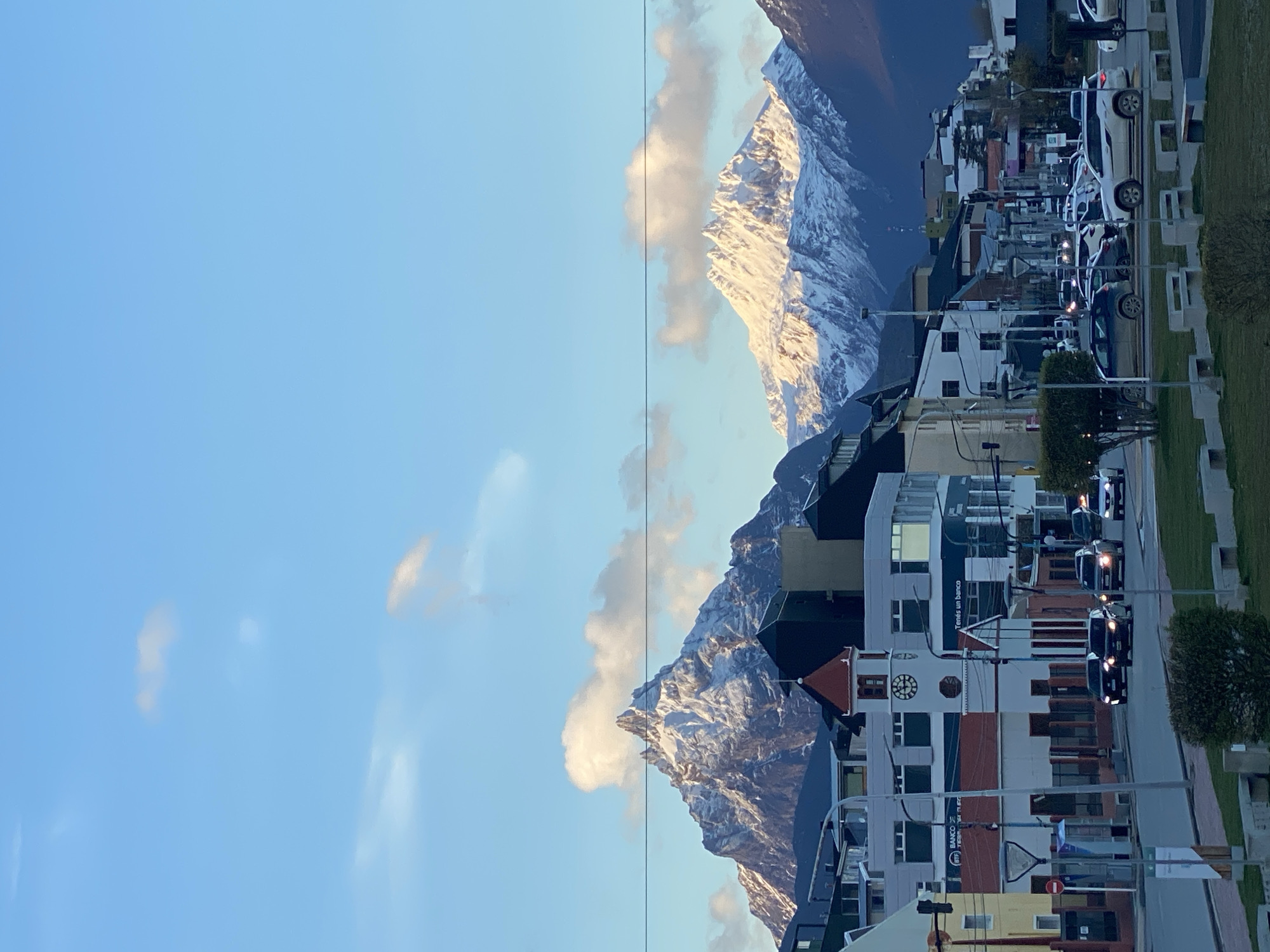The image depicts a street scene in Ushuaia, Argentina, with snow-capped mountains in the background. The sky is blue with some clouds. On the right side of the image are buildings along a street with cars parked and driving. There are streetlights and some greenery. The mountain range appears directly behind the buildings. A power line runs across the horizon between the sky and the mountains.