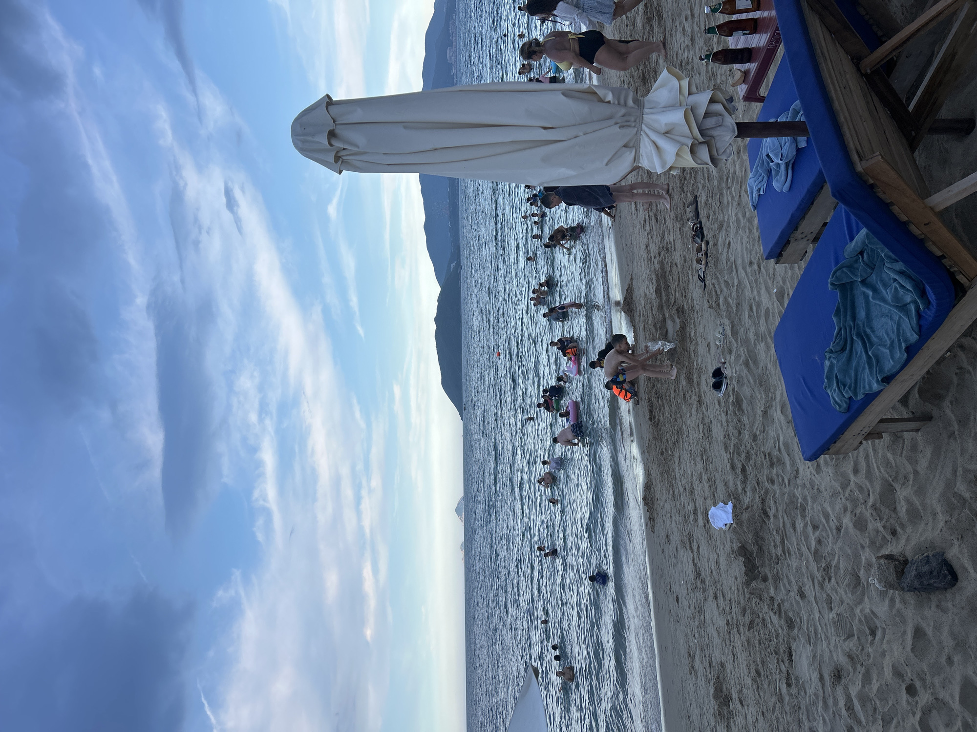The image depicts a beach scene in Nha Trang Ward, Vietnam, during what appears to be late afternoon or early evening, given the sky conditions and lighting. The ocean is filled with numerous people swimming and playing, some using inflatable rings. The beach is sandy, with a few lounge chairs with blue cushions and towels visible in the foreground. A closed beach umbrella stands prominently to the right of the frame. Various items, including a white t-shirt, a pair of flip-flops, and bottles of drinks on a small table, are scattered on the sand. In the background, hills and mountains form the coastline under a partly cloudy sky with shades of blue and grey.
