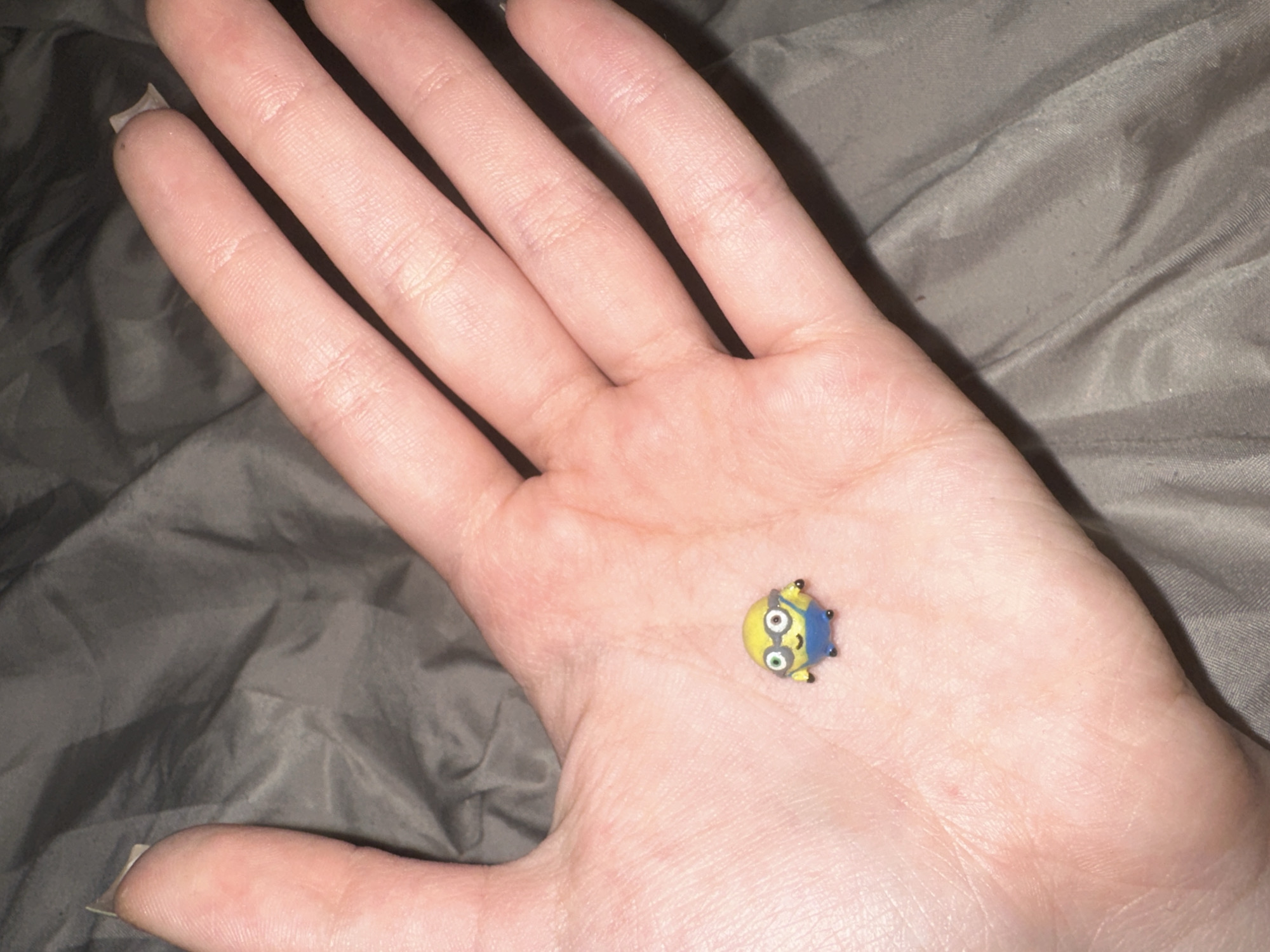 A close-up shot shows a small Minion toy figure resting in the palm of a human hand. The Minion is yellow with blue overalls and is wearing round glasses. The hand is open, and the background consists of a textured gray surface, possibly fabric. The image does not provide any specific location information, nor does it appear to be located in Petropavlovsk-Kamchatsky, Russia.