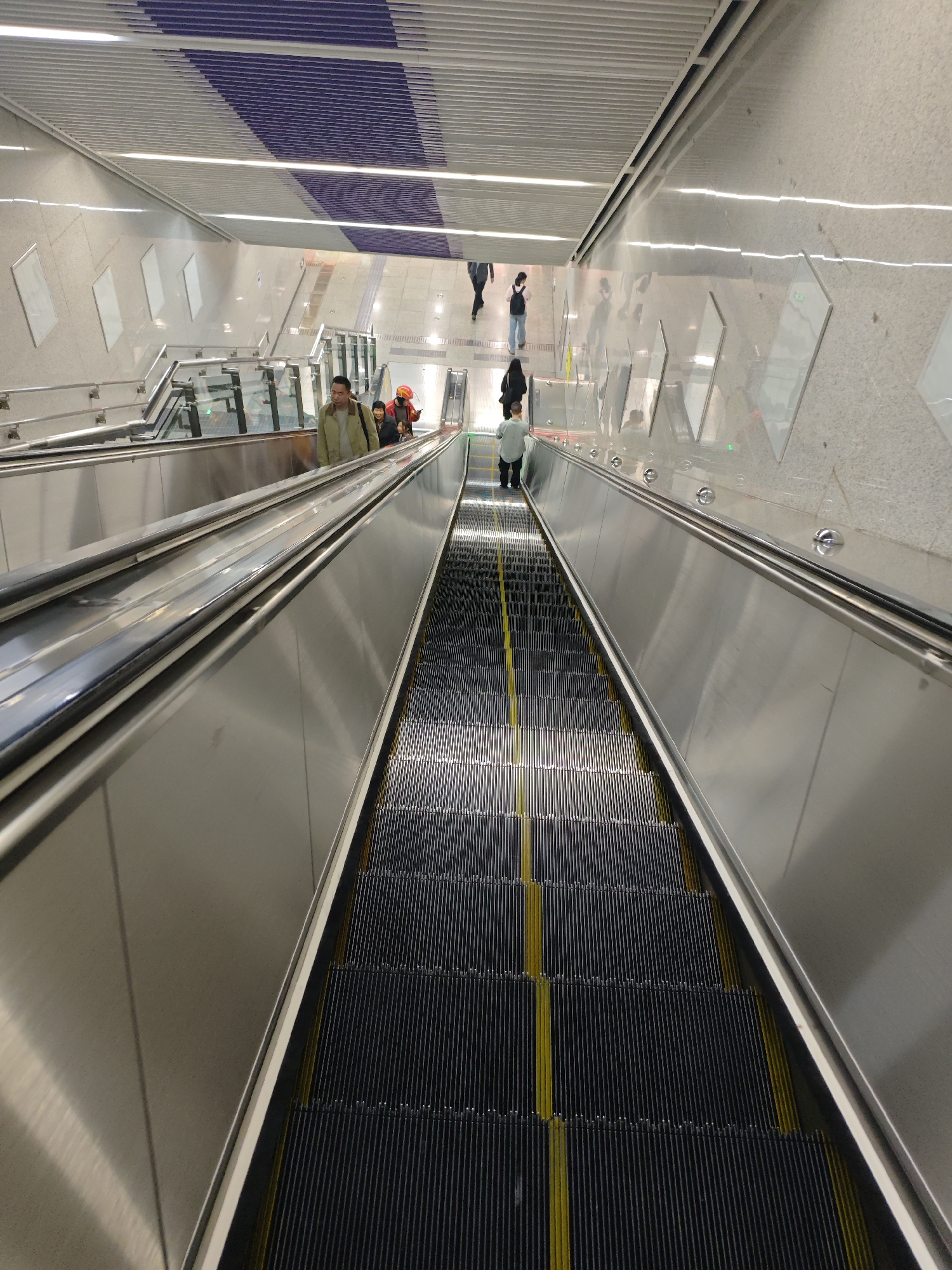 The image shows a descending escalator in what appears to be a modern subway or train station. The escalator has a black tread with yellow lines, and silver railings on both sides. People are on and around the escalator, with some standing on it and others walking at the top. The architecture is modern, featuring metallic wall panels and a ceiling with blue and white patterns. The scene could be in the Liangjiang New Area of Chongqing, China, given the modern infrastructure and subway system development in the city. The image captures a common scene of urban transportation.