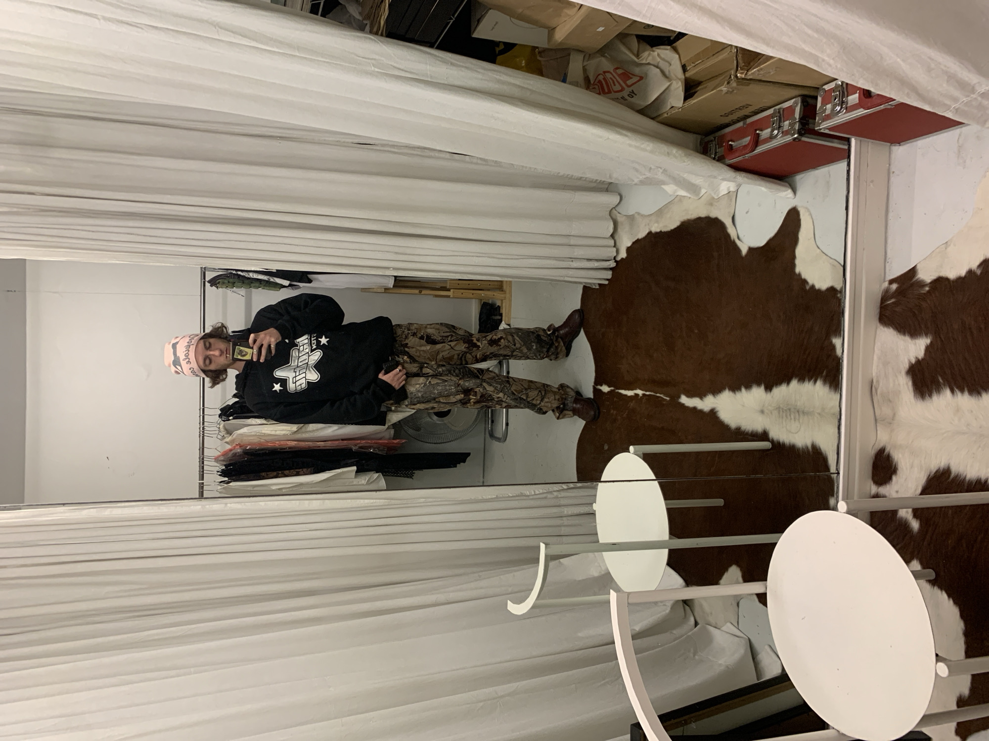 A full-length mirror reflects a person dressed in a black hooded sweatshirt, camouflage pants, and brown shoes. The person is holding a smartphone and wearing a pink and white patterned beanie. The reflection shows a rack of clothes behind the person. The area around the mirror features a white curtain, a cowhide rug, and a white side table with two circular surfaces. Boxes and containers are stored above the mirror. No explicit location or reference to Buenos Aires, Argentina, is identifiable in the image.