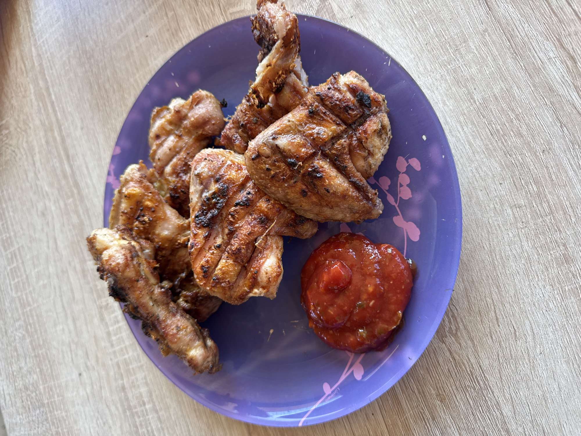 On a light brown wooden surface, a round, purple plastic plate sits holding several grilled chicken wings and a small pile of reddish-orange sauce. The chicken wings exhibit grill marks and some charred areas. The plate features a pattern of stylized pink leaf-like shapes. The location is undetermined, as no information is present to place the image in Chaikovsky, Russia.