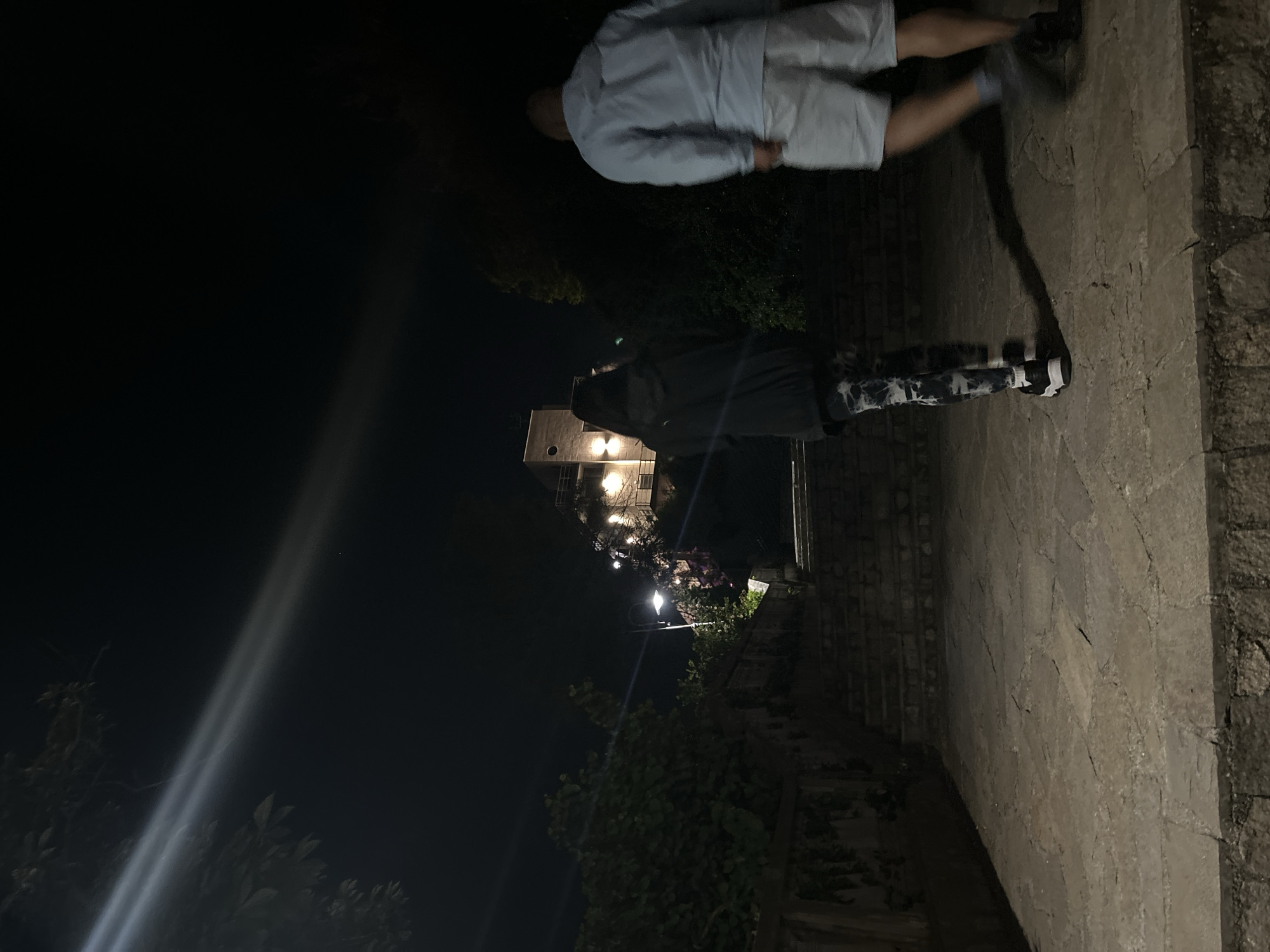 At night, an outdoor scene shows a person in a dark hooded cloak and pants with a black and white pattern standing on a stone walkway. Another person in a white shirt and shorts walks towards the upper right. A building with lit windows stands behind the hooded figure. Streetlights illuminate the area and trees and plants are visible. The location could possibly be in Burgas, Bulgaria.