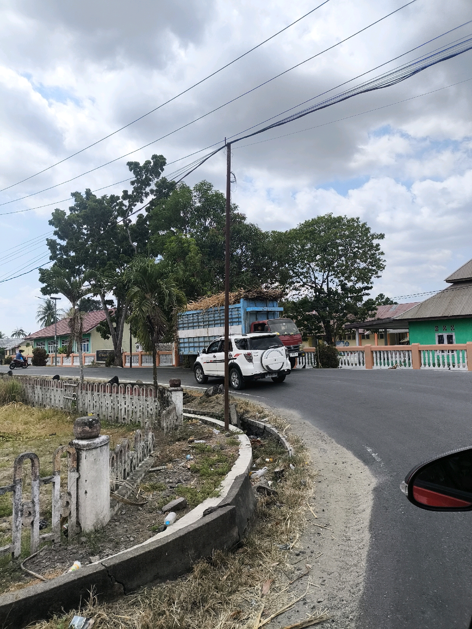 The image depicts a street scene in Tenilo, Indonesia, under a cloudy sky. A white SUV is driving on a paved road, approaching a large blue truck loaded with agricultural produce, likely sugarcane. The truck is red and appears to be older. Both vehicles are moving along a curved road that passes by several buildings. On the left side of the road, there is a white picket fence with concrete posts and some overgrown vegetation. A person on a motorcycle is seen in the distance on the left. The buildings appear to be single-story structures, one painted green and others a light yellow or beige, with some having red tiled roofs. The area alongside the road shows signs of neglect, with debris and dry grass. Power lines are visible stretching across the upper portion of the image. The perspective suggests the photo was taken from another vehicle.