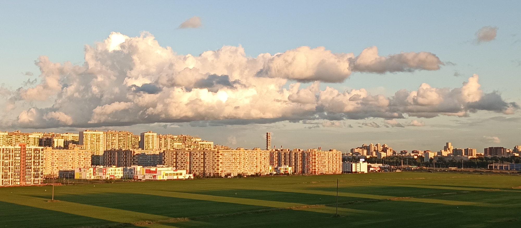 The image captures an aerial panoramic view of a suburban cityscape in Bugry, Russia, under a partly cloudy sky. The foreground is dominated by a vast expanse of vibrantly green grass, casting long shadows from an unseen light source, likely the setting sun. A solitary wooden pole stands vertically in the field, with faint shadows suggesting other indistinct figures.

In the mid-ground, a dense cluster of apartment buildings stretches horizontally across the frame. These buildings are characterized by their uniformity, with many featuring similar architectural styles and color palettes, predominantly light beige and orange. Some buildings exhibit colorful accents, with sections painted in bright red, yellow, and white. A few lower-rise structures, possibly commercial buildings or community centers, are interspersed among the taller residential blocks. One prominent structure in this area is a tall, cylindrical tower with a distinctive ribbed exterior.

The background reveals a more distant, slightly hazy skyline of a city, featuring a variety of building heights and styles, hinting at a more established urban center. The sky above is a pale blue, punctuated by large, cumulus clouds that are illuminated from below, suggesting they are catching the warm light of either sunrise or sunset. The overall impression is one of a developing residential area on the outskirts of a larger city, bathed in the soft light of late afternoon or early evening. There are no visible people or animals in the scene.