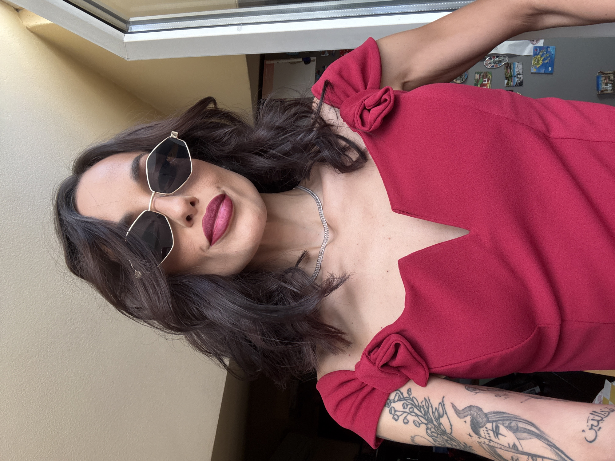 A woman is posing indoors near a window. She is wearing a red dress with bow details on the shoulders, sunglasses, and a silver necklace. She has dark hair and dark lipstick. A portion of a tattoo is visible on her arm. The background includes a wall with pictures and a window. The location is identified only as the city of Pleven, Bulgaria.