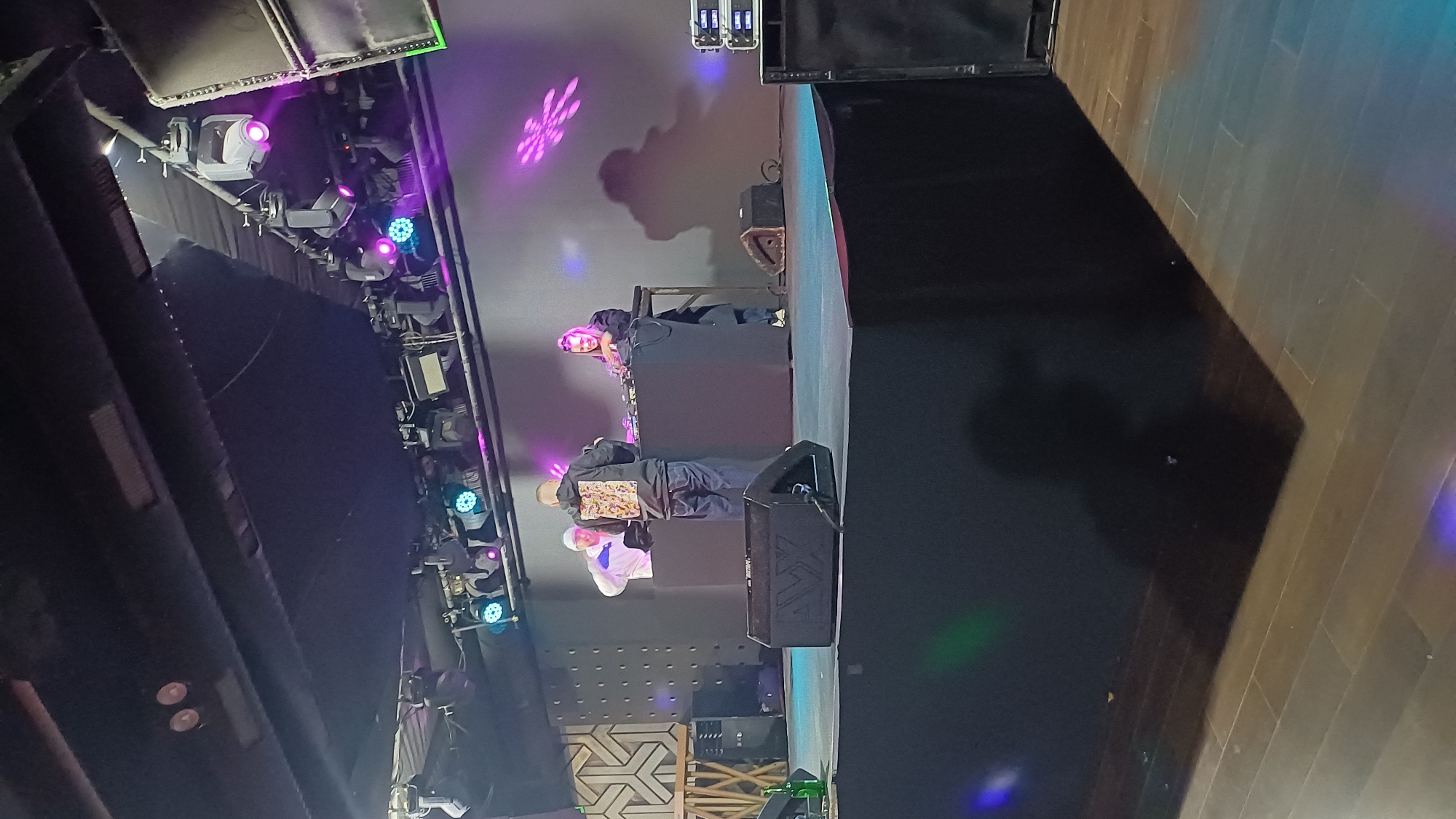 The image shows a stage setup at a venue, possibly in Sofia, Bulgaria, although the specific location is not definitively identified. There are two people visible on the stage near a DJ setup. One person is wearing a light-colored shirt and the other is wearing a dark jacket with an elaborate design on the back. A speaker is positioned prominently on the stage. Stage lighting fixtures and equipment are visible overhead, with purple and blue lights illuminating the scene. There is a dark background and a dark floor that transitions to a lighter, possibly wooden, floor. The image appears to be taken from a low angle, looking up towards the stage.