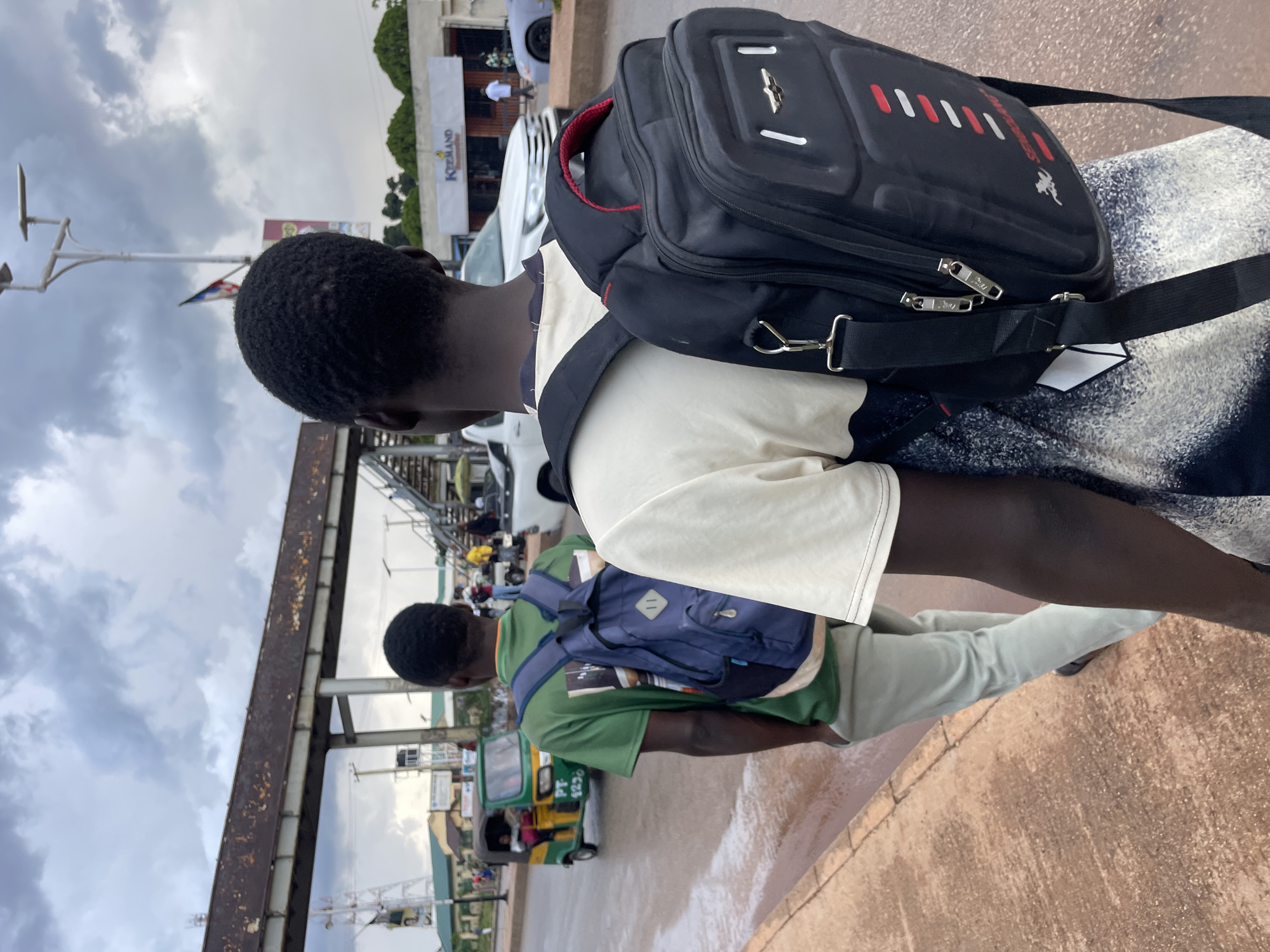 Two young Black men walk on a sidewalk in Jos, Nigeria. The man in the foreground, facing away from the camera, wears a cream-colored t-shirt with a patterned design and a large black backpack. The man behind him, also walking away, wears a green t-shirt and a blue backpack. In the background, a green and yellow auto-rickshaw is visible, as well as cars and a pedestrian overpass. The sky is overcast with dark clouds. The road appears wet, suggesting recent rain. The scene is likely on a street in Jos, a city known for its hilly terrain and mining history.