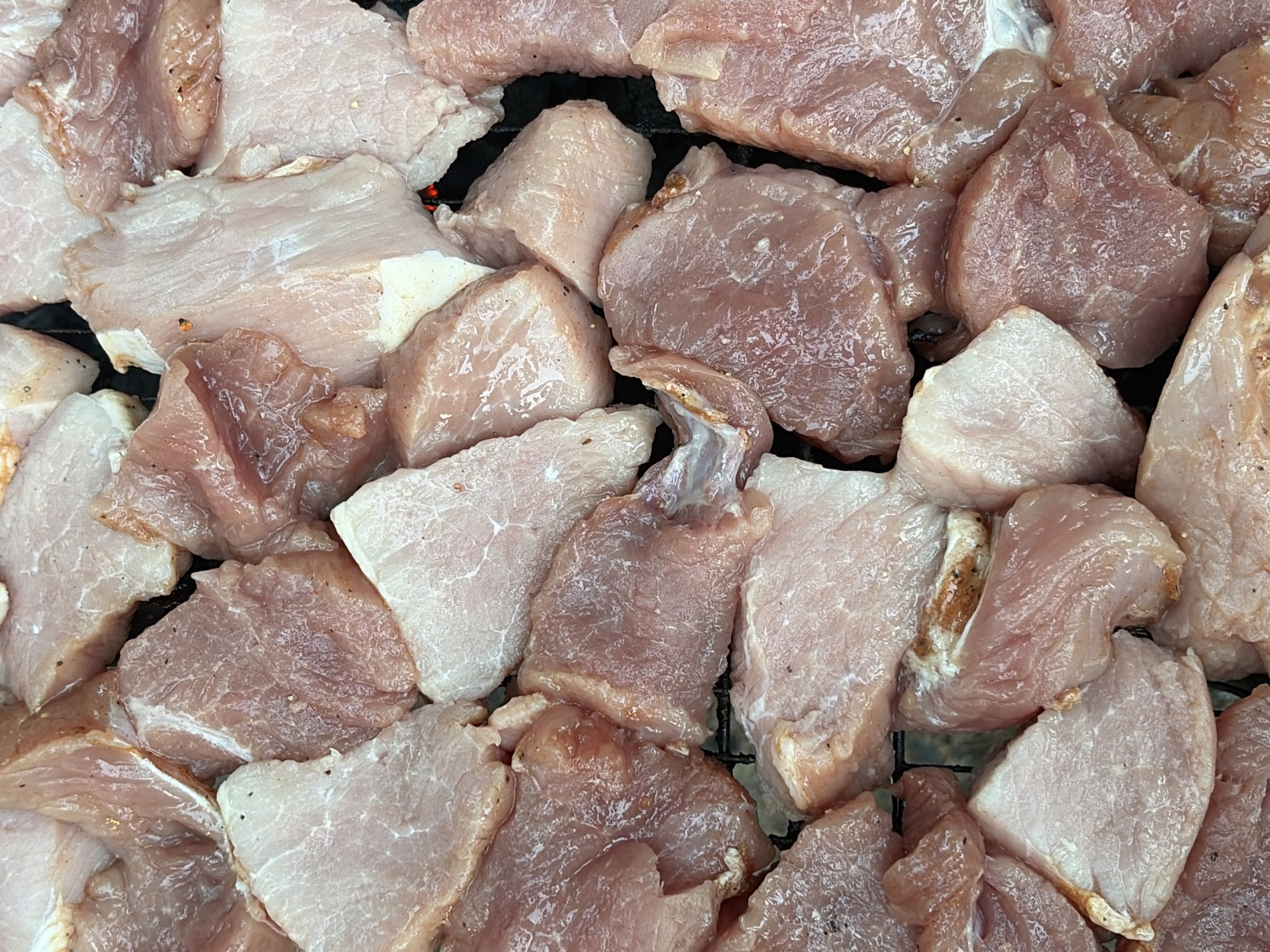 The image shows a close-up, overhead view of numerous raw, cubed pieces of pork meat arranged on a black grill grate. The meat varies in color from pale pink to a deeper reddish-pink, with some pieces appearing slightly translucent and moist. There are small flecks of seasoning, likely pepper, visible on some of the pieces. The background consists of the dark, textured surface of the grill. There are no people or visible actions occurring in the image. The image does not contain any information that would suggest a specific location within the city of Snegiryovka, Russia, or any other geographical context.