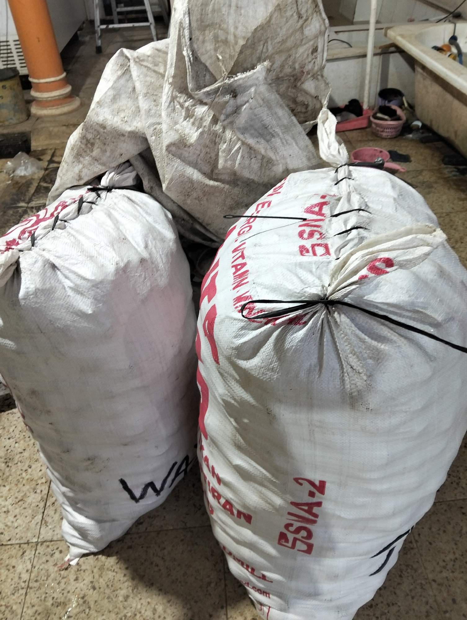 Two large white woven sacks are centrally positioned in the foreground, resting on a tiled floor. The sack on the left has the black marker inscription "WA" visible near its base, partially obscured by the sack's folds. The sack on the right displays red lettering that reads "SSIA-2" and some of what appears to be "TAIN," with additional red text obscured by the sack's closure. Both sacks are cinched shut with black ties. Behind the sacks, a large, weathered, off-white tarp or fabric lies crumpled. Further in the background, a bathtub is visible on the right, containing various items, and a pink basket with more objects. To the left, an orange pipe extends vertically, supported by white rings. The floor appears to be a light-colored tile with a speckled pattern, and there are some scattered water droplets. The overall environment suggests an indoor, possibly utilitarian or storage area in Muncar, Indonesia.