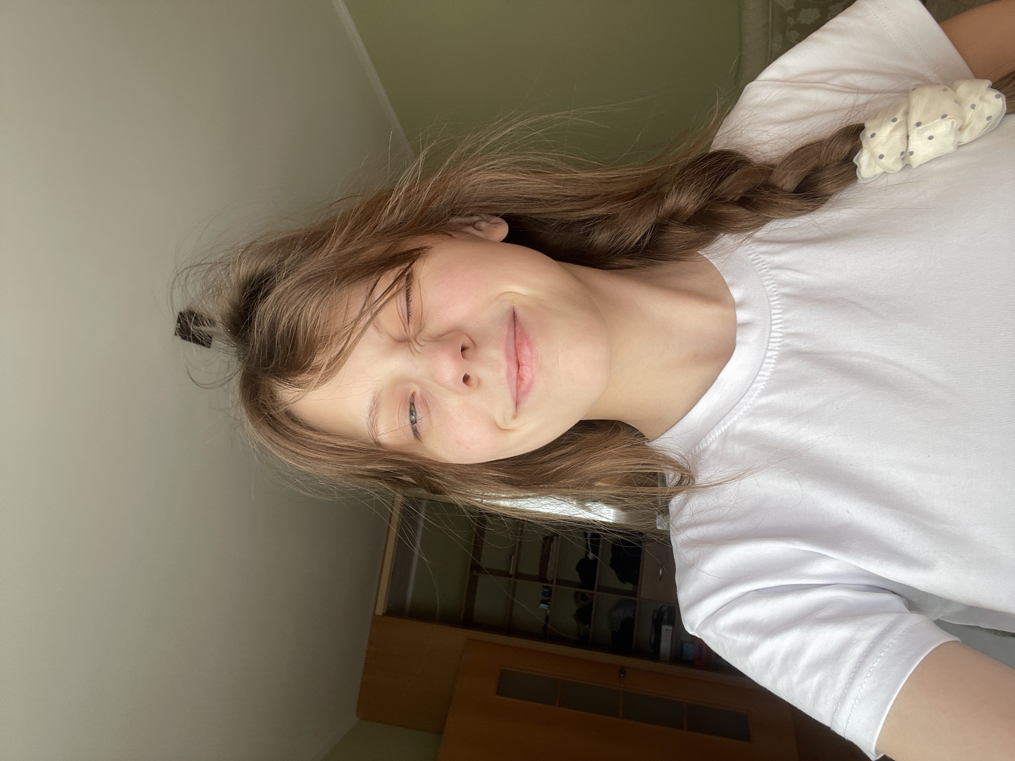 A young Caucasian female is captured in a selfie, facing the camera with her right eye closed in a wink and her left eye partially open. Her lips are slightly pursed in a smile. Her light brown hair is disheveled, with some strands sticking up from her head and a braid with a white polka-dot scrunchie is visible on her right side. She is wearing a plain white t-shirt. Behind her, on the left, is a closet with wooden doors, one slightly ajar, revealing shelves with various items inside. The background also shows a light green wall and a portion of a white ceiling with a light fixture. The setting appears to be an indoor residential environment. No specific location in Ufa, Russia is indicated.