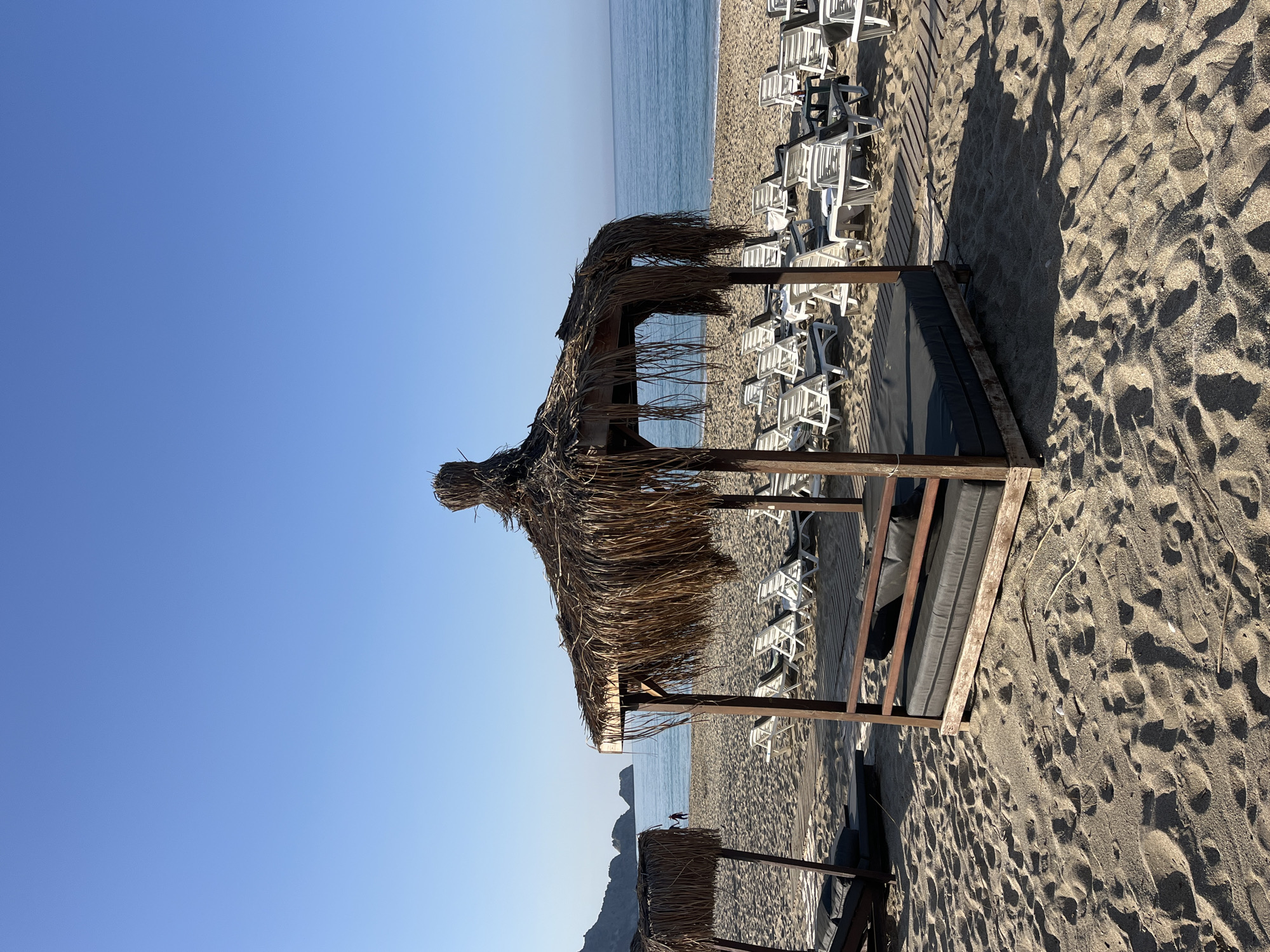 The image shows a beach in Alanya, Turkey. A cabana with a thatched roof stands on the sand, with beach chairs visible in the background. The sea is visible in the distance under a clear blue sky. The sand shows patterns of wind and footsteps. There is a mountain visible in the horizon, to the left side of the image.