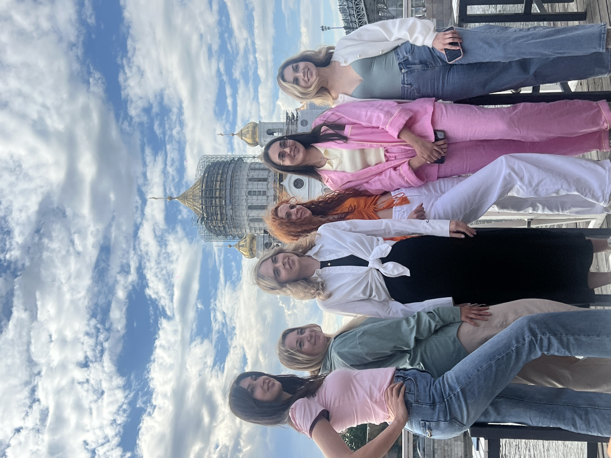 Six women of varying ethnicities stand in a row, one behind the other, posing for a photograph. The background features a partially constructed white cathedral with golden domes, scaffolding, and a backdrop of blue sky with white clouds. The setting is the city of Moscow, Russia, as evidenced by the presence of the cathedral. The women are dressed in casual, contemporary clothing, including jeans, blouses, dresses, and sweaters. One of the women is holding a smartphone. They are standing on a structure with a metal framework.