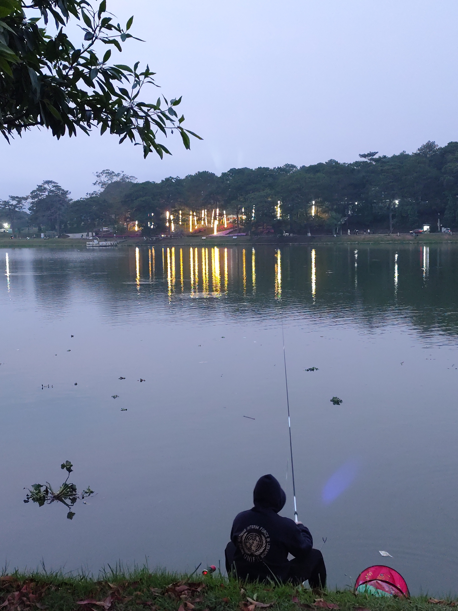 An individual is fishing at Xuan Huong Lake in Da Lat, Vietnam. The person, wearing a dark hooded sweatshirt and dark pants, is seated on the grassy bank of the lake, holding a fishing rod. A tackle box is next to them. The lake reflects light from buildings or decorations on the opposite bank. The background consists of trees and a cloudy sky. Some aquatic plants are visible in the lake.