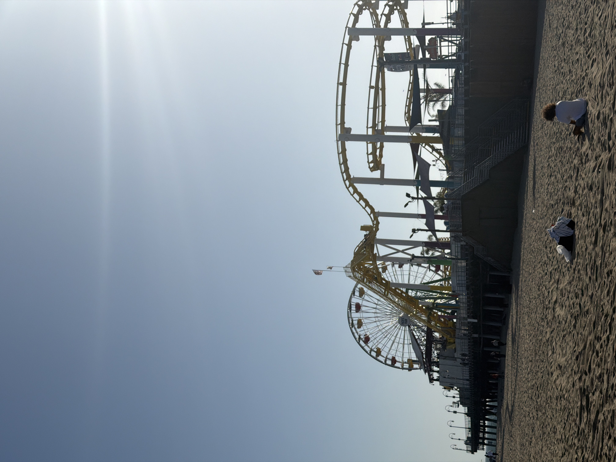This outdoor, rotated, daytime shot features the Santa Monica Pier with its Ferris wheel and roller coaster. The pier structure is dark, with visible supports extending down into the sand. A Ferris wheel, with yellow and white spokes and gondolas is visible toward the bottom-center of the image. A roller coaster, also constructed with yellow tracks and white supports, runs along the top of the pier. The sky is a light blue color, with the sun in the upper-left corner of the frame. The beach occupies the right side of the image and shows a sandy surface. There are two people on the beach to the right of the pier.