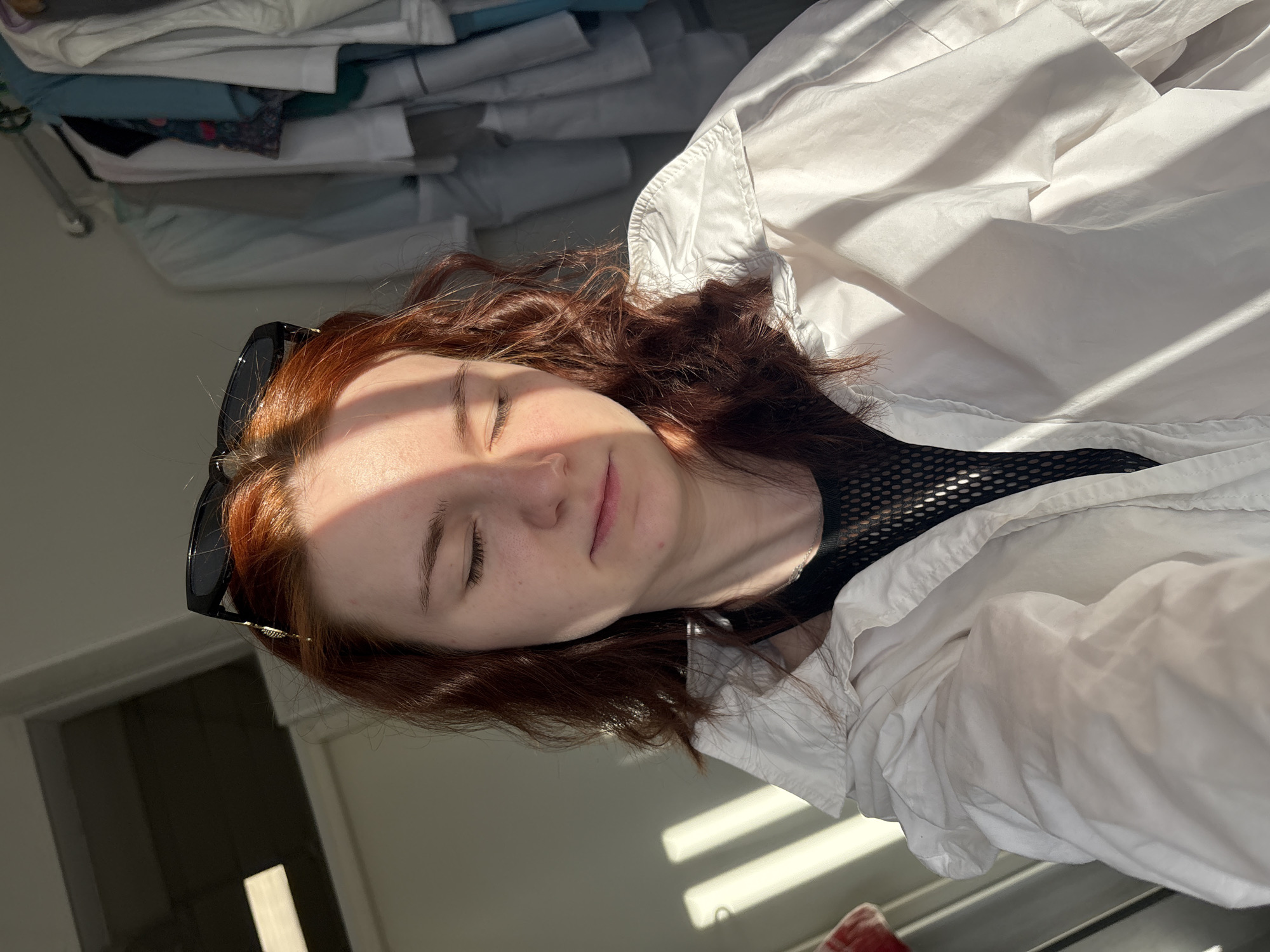 A close-up, angled photograph captures a young individual, likely female, with their eyes closed, basking in natural sunlight. The subject has fair skin and distinct reddish-brown, wavy hair, with black framed sunglasses pushed up onto their head. They are wearing a white collared jacket or shirt over a black, mesh-patterned top.
Strong, parallel streaks of sunlight and shadow diagonally illuminate the scene, falling across their face, hair, and clothing, suggesting a sunny day, possibly morning or late afternoon, with light streaming through a window or blinds. The person's expression appears peaceful and relaxed.
In the blurred background to the upper left, stacks of folded light-colored clothing or textiles are visible, hinting at an indoor setting such as a bedroom or living space. The overall atmosphere conveys a moment of calm and warmth. There is no visible text. The location is an unknown indoor setting in Russia.