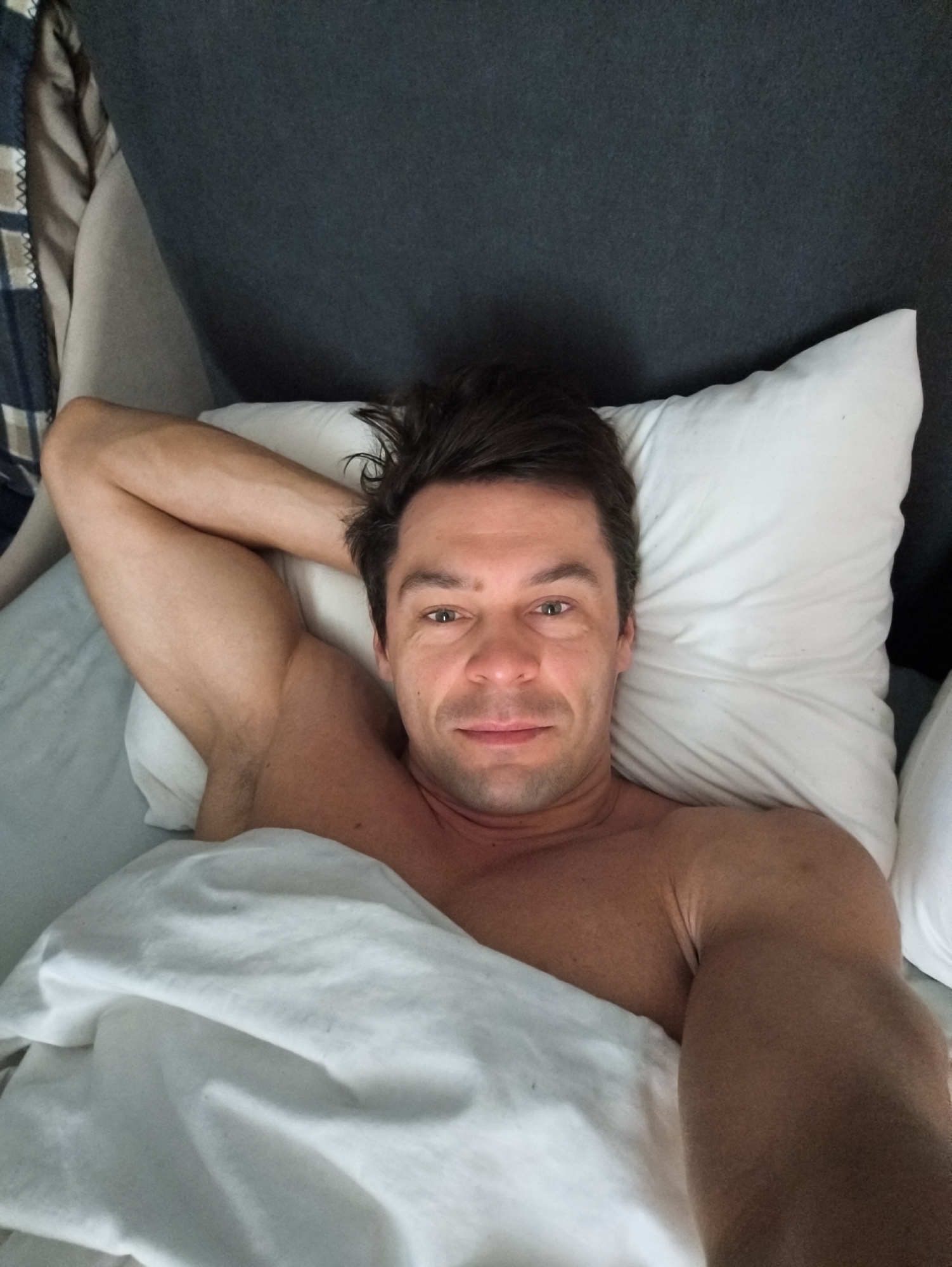 A man is lying in a bed with white sheets and a white pillow. His head is resting on the pillow and he has one arm raised behind his head. He is shirtless and partially covered by the sheets. The headboard of the bed is a dark color. A plaid blanket is visible on the left side of the image. The location cannot be determined from the image.