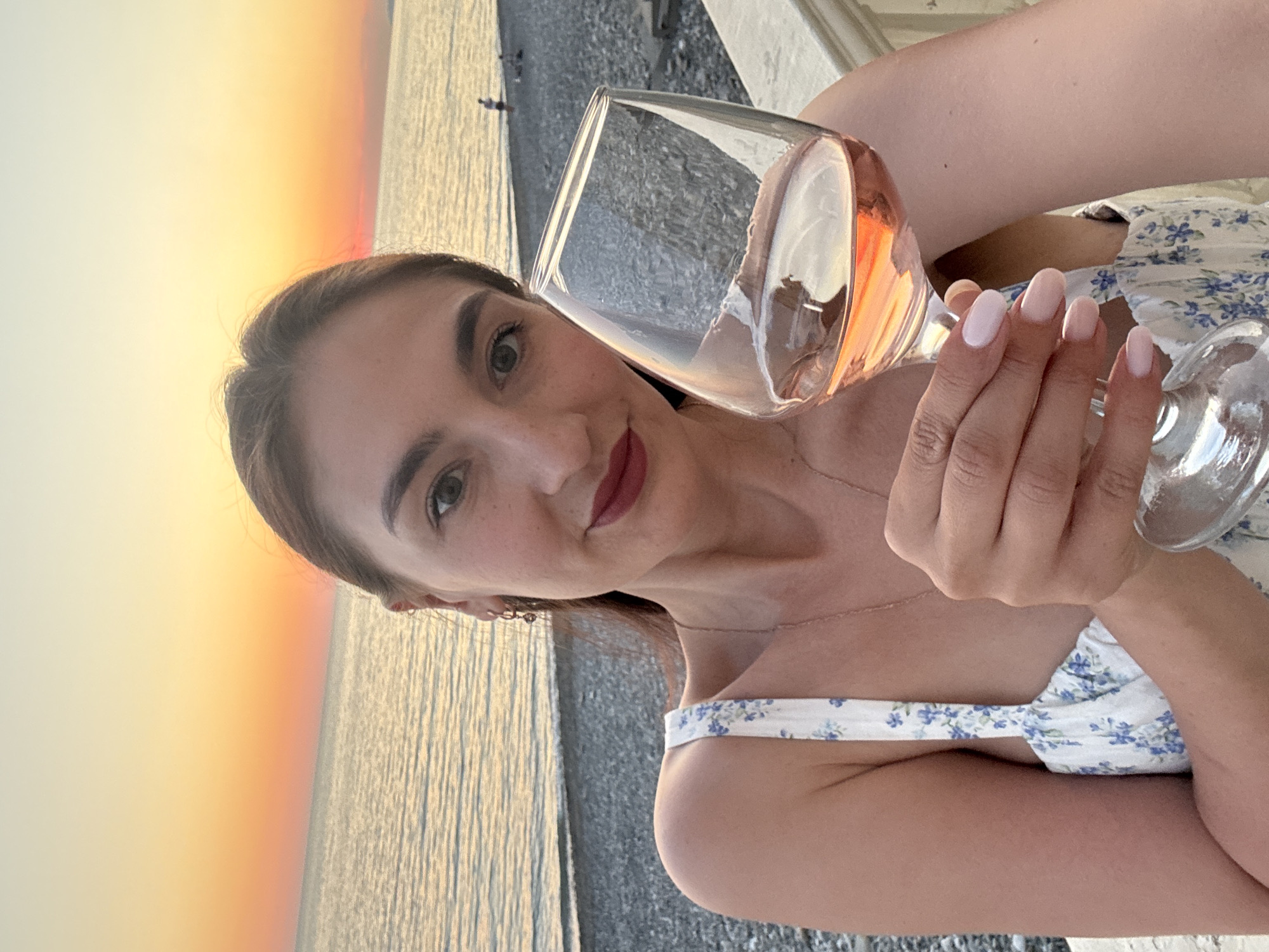 The image captures a young adult female in a selfie during sunset at a location identified as Sochi, Russia. The woman, positioned in the foreground, has brown hair, brown eyes, visible freckles, and wears dark red lipstick. She is dressed in a white, blue floral-patterned sundress, with a thin gold chain necklace and small hoop earrings. Her fingernails are painted a light pink/nude color. She holds a stemmed glass containing rosé wine in her right hand, positioned slightly forward, and looks directly at the camera with a slight smile. The background features a calm sea extending to a clear horizon under a sunset sky, which displays vibrant gradients of orange and yellow hues. A pebble beach lines the shore, and in the distance, two indistinct human figures are visible on the beach. A white stone railing or parapet is partially visible in the lower right, indicating the woman is on an elevated platform overlooking the beach and sea.