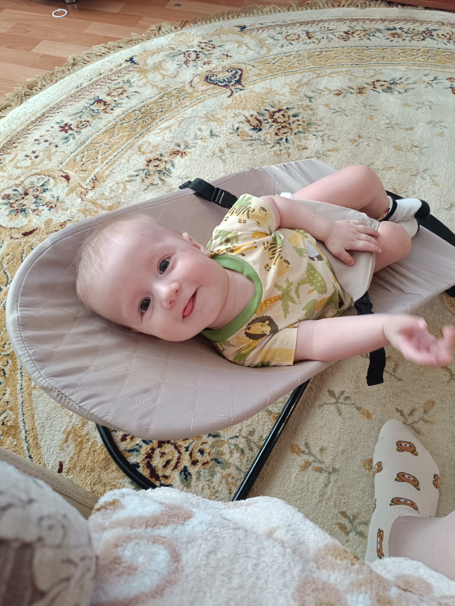 A baby in a beige baby bouncer sits on an ornate, beige, round rug. The baby, wearing a yellow and green onesie and white socks, is looking at the camera with a small smile. The baby's tongue is slightly sticking out. Part of a foot in a sock with cartoon bears is visible in the foreground, covered in a white and beige patterned blanket. The location is indoors on what appears to be light-colored wood flooring. There is no verifiable information to suggest this scene is located in the city of Tobolsk, Russia.