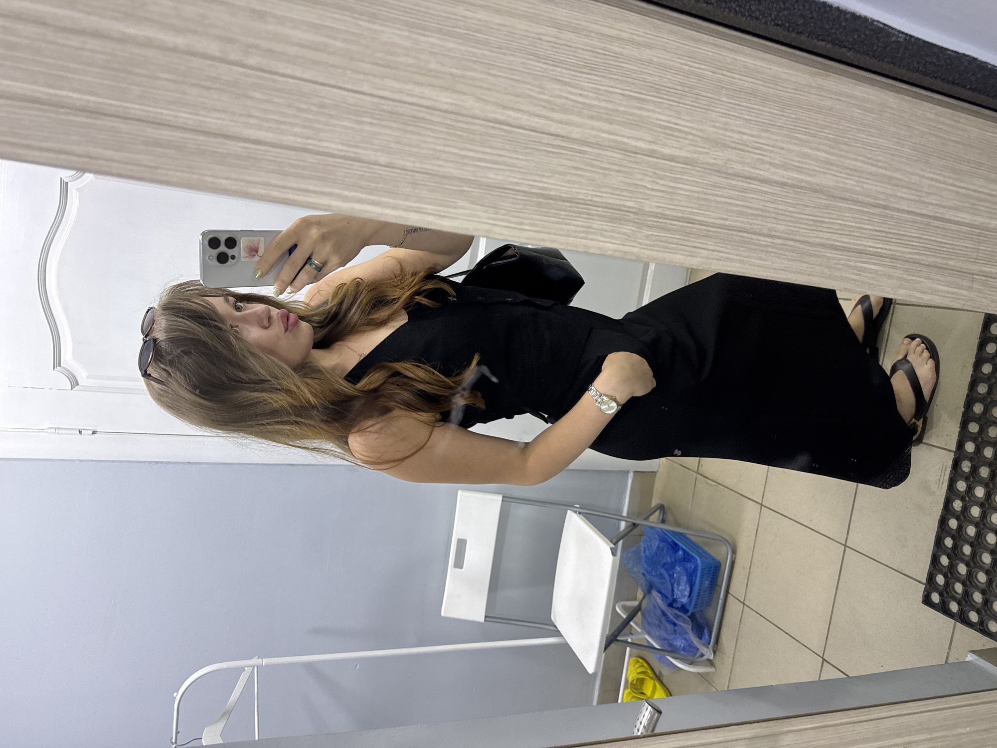 A young woman with long, wavy brown hair, wearing sunglasses on her head, a black sleeveless dress, a watch, and black sandals, stands in a room and takes a selfie in a mirror. She is holding a smartphone in her right hand. To her left, a white folding chair is visible with blue plastic bags on it. Further to the left, a white clothing rack with a single hanger is present. The flooring is tiled. The background consists of a light blue wall and a white doorframe. The room appears to be an indoor space, possibly a dressing room or a hallway in a residential building or a public facility. The image does not contain any specific landmarks or identifiers that would pinpoint its location to Chaikovsky, Russia.