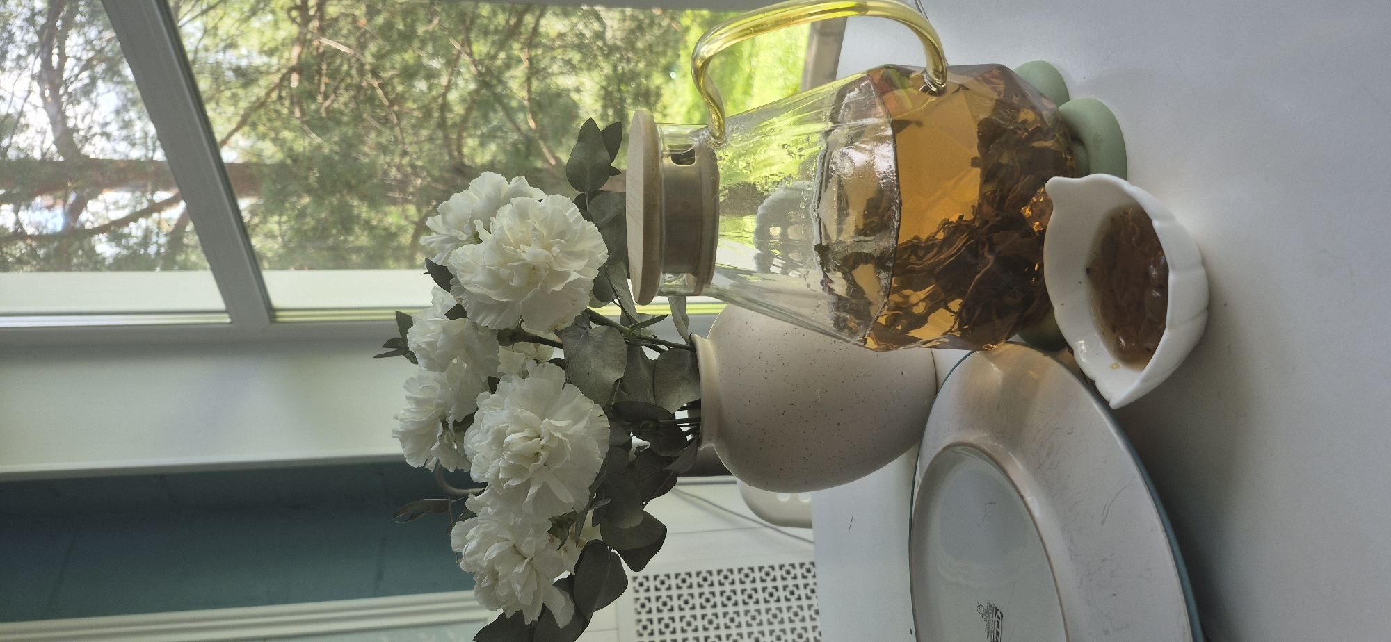 The image depicts a close-up, eye-level view of a table set for tea, with a window in the background. The table is white, and on it, to the left, is a white plate with a light blue rim. Next to the plate, there is a small white, leaf-shaped dish containing a brownish jam. In the center of the image, a clear glass teapot filled with amber-colored tea and visible tea leaves sits on a green coaster. The teapot has a distinctive geometric shape and a gold-colored handle. Behind the teapot, a white ceramic vase holds a bouquet of white flowers with green stems and leaves. The window in the background reveals a blurry view of green trees and foliage under natural daylight, suggesting an outdoor environment. The overall scene evokes a peaceful, domestic setting. There are no people or animals present in the image. The location is specified as Bezvodnoye, Russia.