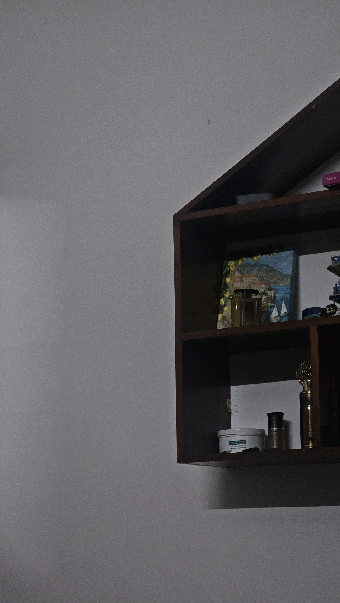 A dark brown, angled shelf is mounted on a light gray wall. The shelf is divided into three sections. The top shelf contains a dark rectangular object. The middle shelf displays a small painting of a coastal town with sailboats, two bottles of perfume, and a blue object. The bottom shelf holds a white jar with a blue lid, a dark bottle, and a golden decorative item. The image is taken from a slightly upward angle, and the lighting casts shadows on the wall and shelf. The scene appears to be indoors, likely a residential setting, with no people present. The location of Choozhattukotta, India, is not discernible from the visual information provided.
