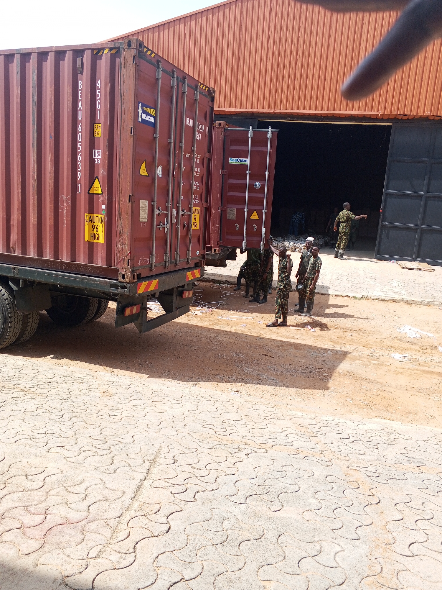 The scene depicts a loading dock at a warehouse in Pasere, Nigeria. Two shipping containers mounted on trucks are positioned in front of an open bay door. Several individuals in military-style camouflage uniforms are present; some appear to be assisting with the unloading process. The environment includes a paved area with a geometric pattern and an orange corrugated metal warehouse structure. The containers are marked with shipping information, including codes and warnings about height and weight. The sky is not visible in the image.