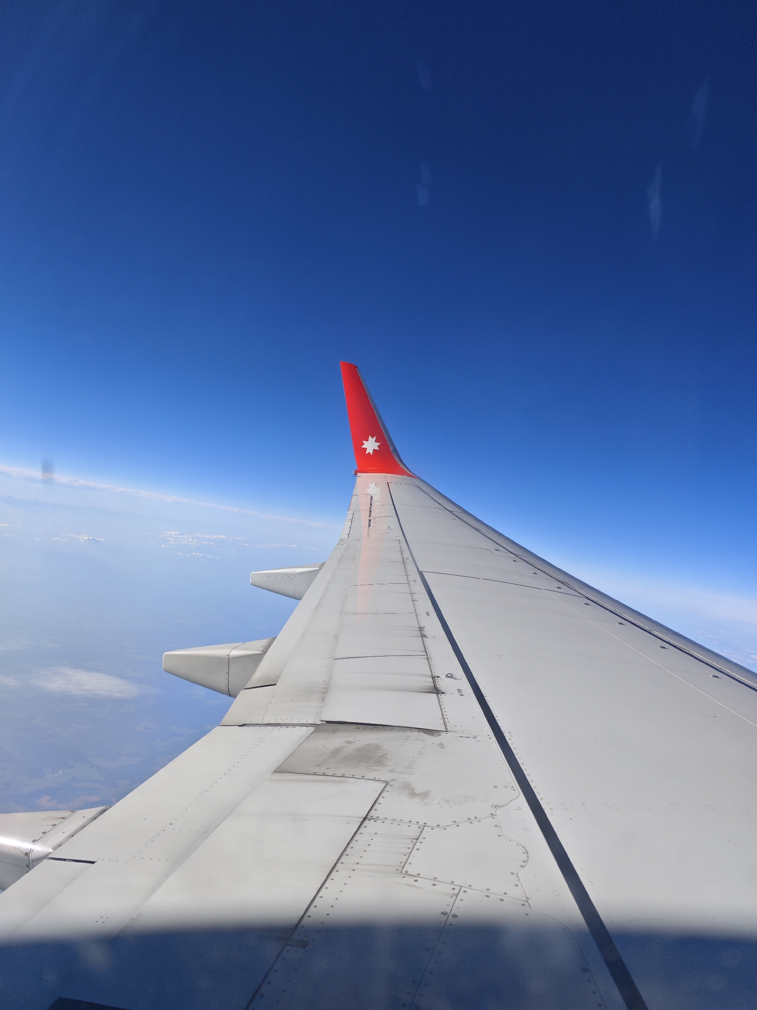 An aerial, wide shot captures an airplane wing and the vast blue sky from the perspective of a passenger. The wing, primarily white, is ribbed and riveted, with a red tip featuring a white star symbol. Below the wing, the Earth is visible, with a hazy layer of clouds and a patchwork of green and brown terrain. The scene is set against a deep, cloudless blue sky.