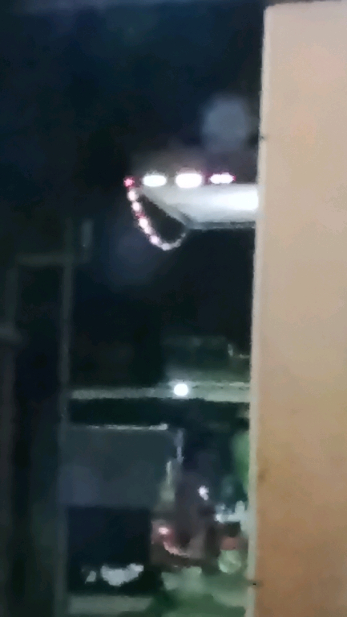 The image, taken at night, shows a blurry exterior view that may be in Ujungrusi, Indonesia. A brightly lit, white awning or overhang is visible in the upper portion of the frame, adorned with a string of small, colorful lights. Below the awning, the scene is dark and indistinct, with hints of structures and possibly vehicles. A tall, light-colored vertical element, possibly a wall or pillar, occupies the right side of the image. The overall impression is of an outdoor area at night, illuminated by artificial light sources.
