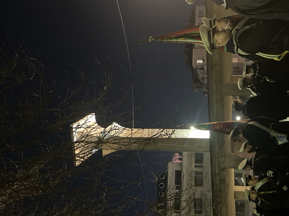 The image depicts a nighttime scene at the Pantheon in the Sea Garden (Morska Gradina) of Varna, Bulgaria. A tall, pale-colored, modernist monument, resembling a vertical pylon with a broader upper section, dominates the left side of the frame, illuminated from its base, casting light upwards. Bare tree branches partially obscure the monument's upper portion and frame the top of the image.
In the foreground and midground, a group of approximately 10-20 individuals, predominantly male, are gathered. They are dressed in dark, formal naval-style uniforms, featuring dark jackets, white peaked caps (some with gold insignia), and some individuals wear sashes (e.g., one visible with blue and yellow stripes). They stand in formation or as a group, appearing attentive. Amongst them, at least two ceremonial flags or banners are held upright, displaying prominent red and green colors, possibly with gold finials at their peaks, partially illuminated by ambient light.
In the background, behind the monument and the gathering, a multi-story building with the Cyrillic letters "AРСОС" visible on its facade is situated to the left. Further back, several other buildings, including what appear to be residential or commercial structures with illuminated windows, are discernible against the dark night sky. A power line or cable runs horizontally across the upper mid-ground. The overall scene suggests a formal, possibly commemorative, gathering or ceremony.