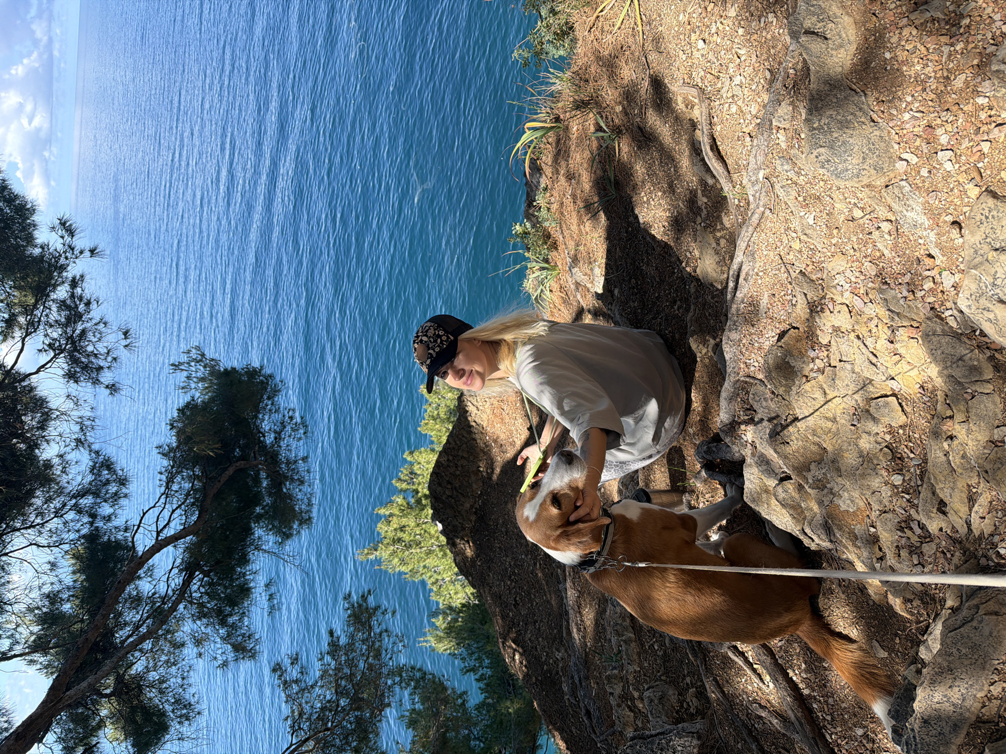 A woman wearing a cap sits next to a brown and white dog on a rocky surface in Petrovac centar, Montenegro. The dog is on a leash. The blue sea is visible in the background to the left, with a tree line at the bottom of the frame. The scene is lit by bright sunlight.