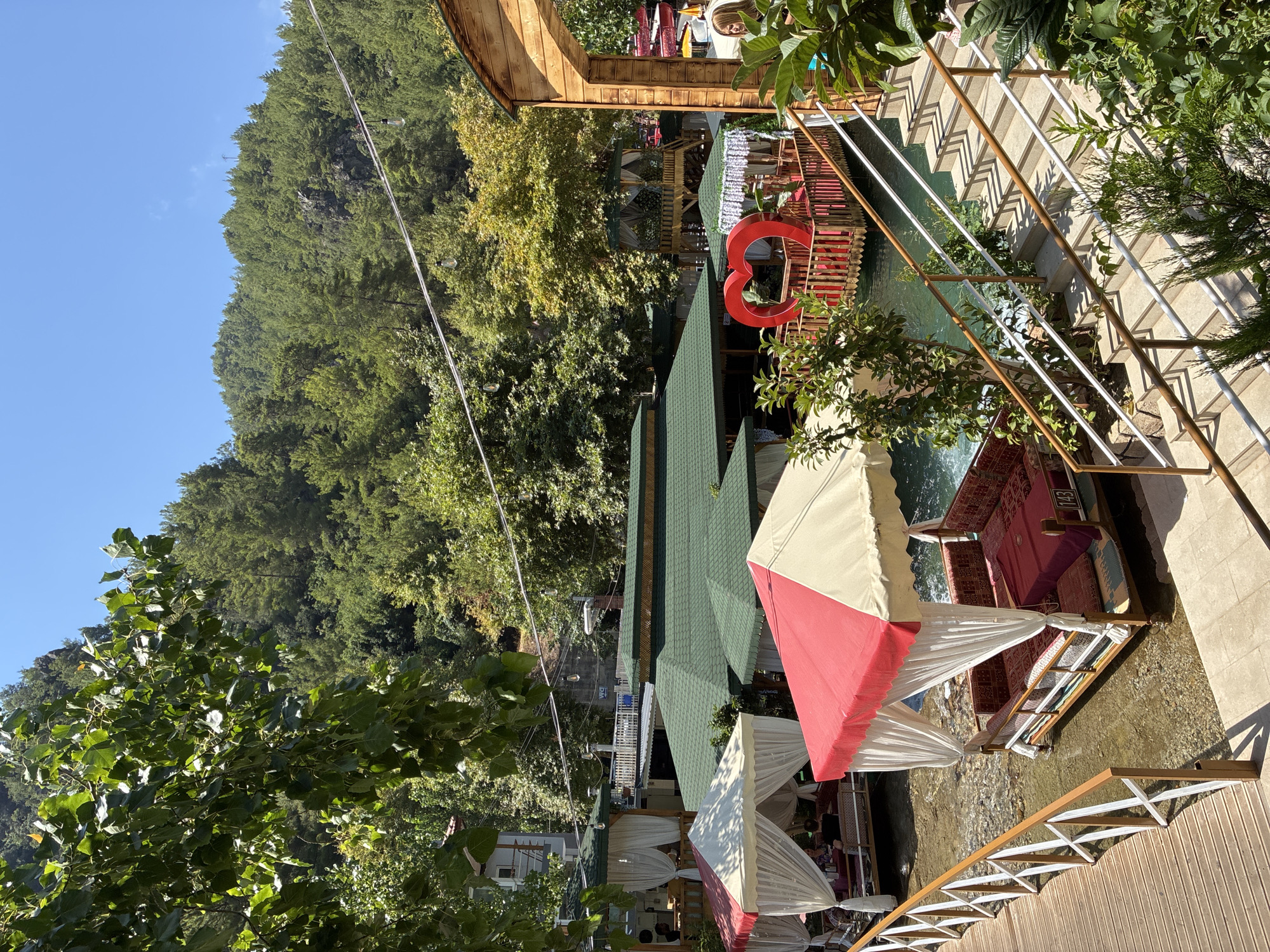 The image depicts a scenic outdoor dining area situated in a natural environment, likely a river or stream, with lush green trees and a hill in the background. The scene appears to be a restaurant or resort with multiple covered seating areas, some of which are partially submerged in the clear water.
In the foreground, a wooden boardwalk leads to an elevated dining platform. Adjacent to the boardwalk, a dining pavilion with a red and white striped canopy covers a seating area. This pavilion features upholstered benches and a table draped with a red cloth. The water beneath the pavilion is shallow, revealing the stony riverbed.
Further into the scene, several other dining pavilions with white canopies are visible, some also situated in or over the water. A prominent red heart-shaped structure is positioned on a wooden platform in the water, surrounded by a red railing. Behind these structures, a larger building with a green roof is visible, along with other trees and foliage.
On the right side of the image, a concrete staircase with wooden and white railings leads down towards the dining area. A woman with blonde hair is standing on the stairs, looking towards the scene.
The overall environment is tranquil and picturesque, suggesting a place designed for relaxation and dining amidst nature. While the location is stated to be Alanya, Turkey, this specific detail cannot be visually confirmed from the image alone.