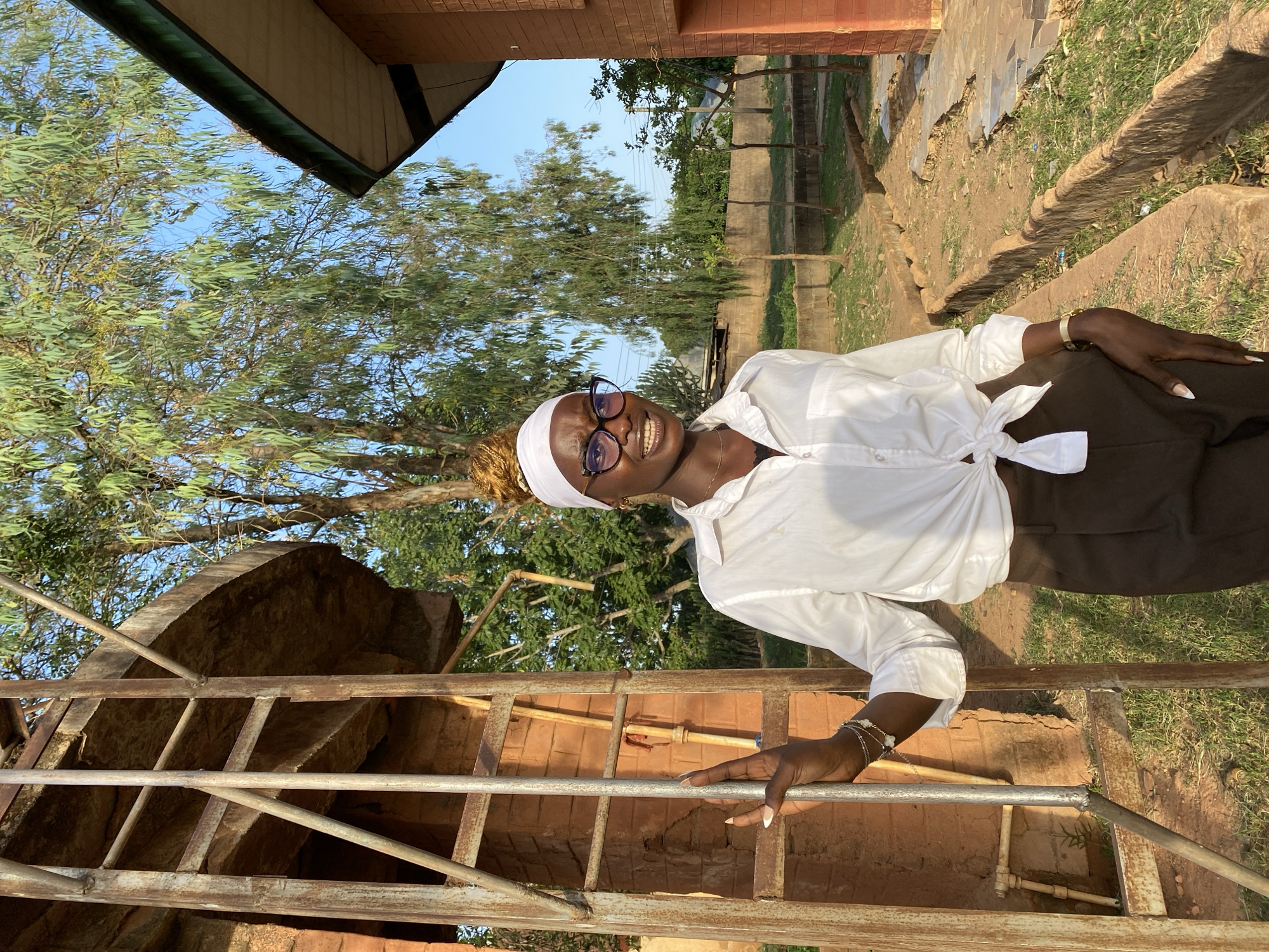 A young woman stands on a metal structure outdoors in what appears to be Vom, Nigeria, based on the landscape and architecture. She is wearing a white shirt tied at the waist, dark pants, a white head covering, and eyeglasses. She is smiling and looking toward the camera. The metal structure is rusty and appears to be part of a construction or renovation project. Behind her are trees, grass, and brick buildings. The environment suggests a rural or semi-rural setting, possibly an academic institution or residential area in Vom.