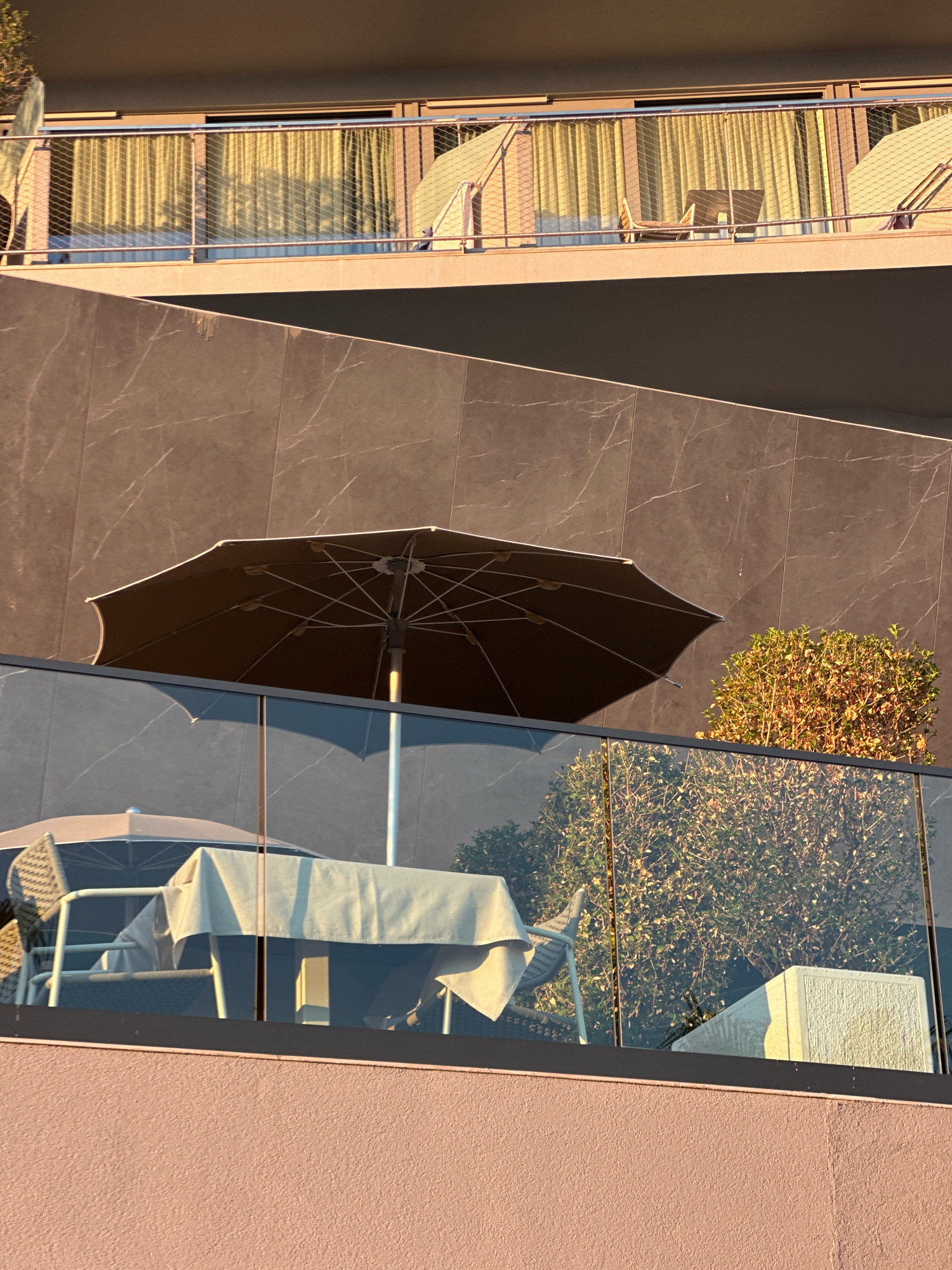 The image shows a close-up view of an outdoor seating area with a table and chairs, covered by an umbrella, on a balcony or terrace. The seating area is behind a glass railing. In the background, there is a building with windows and balconies, and some greenery. The lighting suggests it is either early morning or late afternoon. The scene is located in Rafailovići, Montenegro.