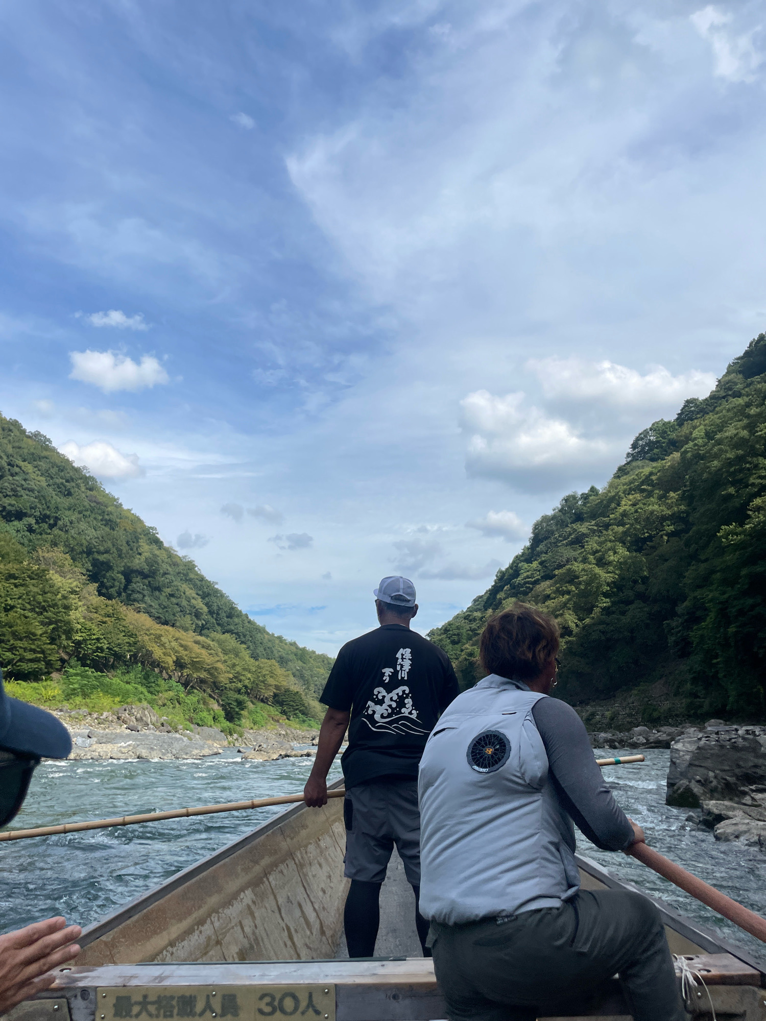 The image shows a group of people on a traditional boat, likely a Hozugawa River Boat. The boat is moving down a river, with lush green mountains flanking both sides. The sky is partly cloudy with a bright blue expanse.
In the foreground, two individuals are visible from the back, seated in the boat. The person on the left is wearing a light grey vest over a grey long-sleeved shirt and dark pants. This person is holding an oar, steering the boat. The person on the right is wearing a black t-shirt with a white design and grey shorts. This person is also holding an oar and appears to be assisting with the navigation. A third person's hand is visible reaching out from the left side of the boat.
The river is wide and appears to have a moderate current, with visible rocks and rapids in some areas. The banks of the river are lined with dense vegetation and rocky terrain. In the lower part of the boat, there is a sign with Japanese characters, and the number "30" is prominently displayed, likely indicating the boat's capacity. The scene suggests a scenic river tour in a natural setting.