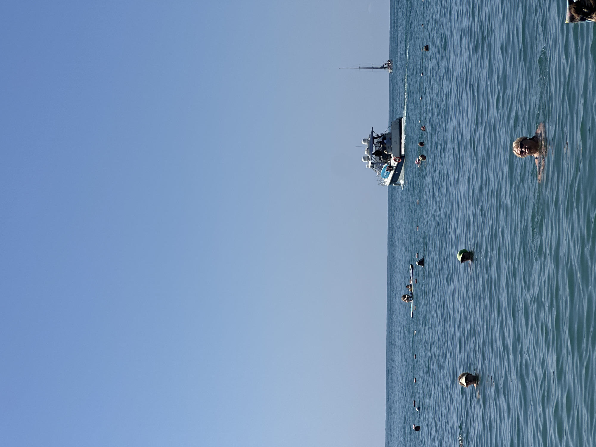 This is a medium-shot, full-frame photograph taken on a sunny day at a beach in Sochi, Russia. The image depicts a clear, bright blue sky meeting a calm, blue sea. In the foreground, a blonde-haired woman with sunglasses is swimming in the water, looking towards the viewer. To her left, another person wearing a yellow cap is also swimming. Further back, in the middle ground, several people are visible in the water, with some appearing to be on a paddleboard. A motorboat with several passengers is cruising in the distance, leaving a wake behind it. To the right of the motorboat, a sailboat with a tall mast is also visible. A line of buoys stretches across the water in the middle ground, marking off a swimming area. The overall scene conveys a relaxed atmosphere of a typical summer day at the beach.