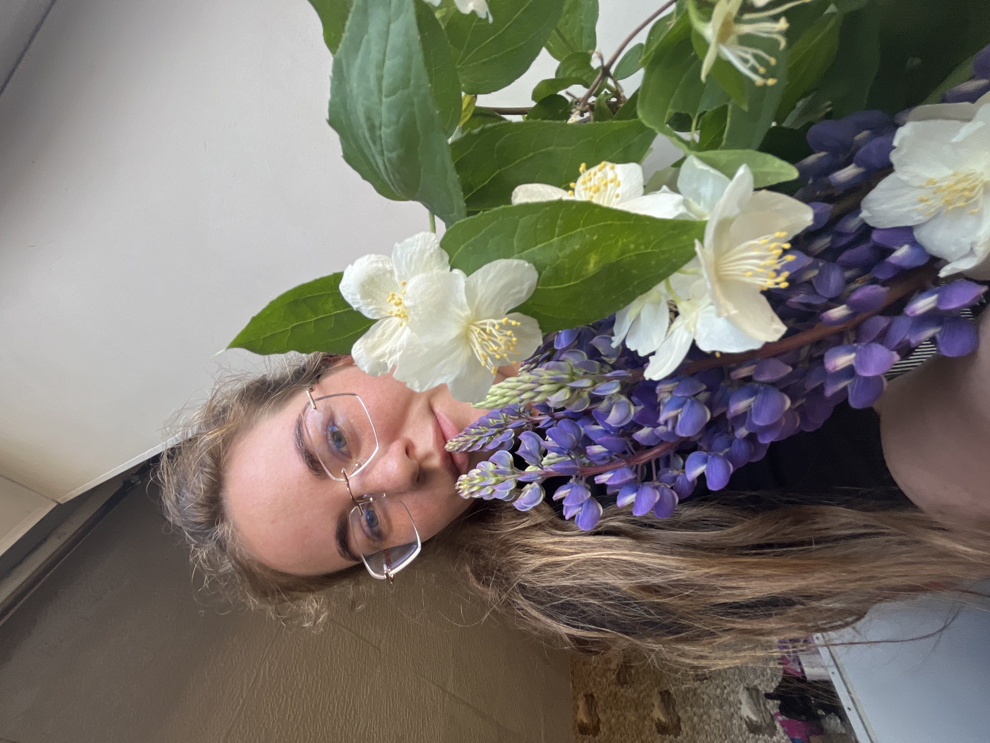 A young adult female, with long wavy light-brown hair and wearing thin gold-framed glasses, is captured in a close-up, upward-angled self-portrait. She looks directly at the camera with a subtle expression. In her hands, she holds a mixed bouquet of flowers comprising multiple stalks of vibrant purple lupine and several clusters of white, five-petaled flowers with prominent yellow stamens, resembling jasmine, accompanied by dark green foliage. The background on the left features a textured, light brown wall with a darker overhead architectural element, possibly a beam or door frame. The background on the right is a plain, light grey wall. The scene is set in an indoor or semi-enclosed environment in the city of Сосново, Russia.
