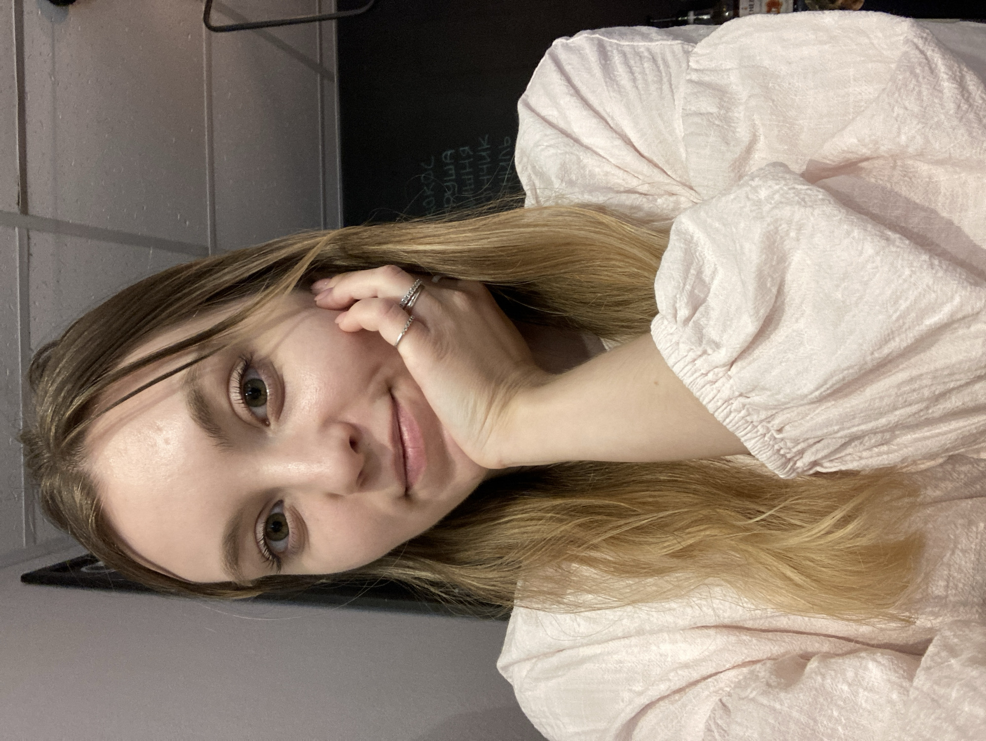 The media file is a portrait of a young woman, shot vertically. She is indoors, with a dark surface behind her that appears to be a chalkboard with some handwritten text. To her left, a grey tiled wall is visible. She has fair skin, light brown hair, and green eyes. Her head is tilted, and her right hand is resting on her cheek, with two rings on her fingers. She is wearing a light pink textured blouse with gathered sleeves. The lighting suggests it might be evening or indoors. The text on the chalkboard is in Russian and includes words such as "любовь," "боль," and "страх." The woman's expression is calm and slightly introspective. The overall mood of the image is intimate and personal.
