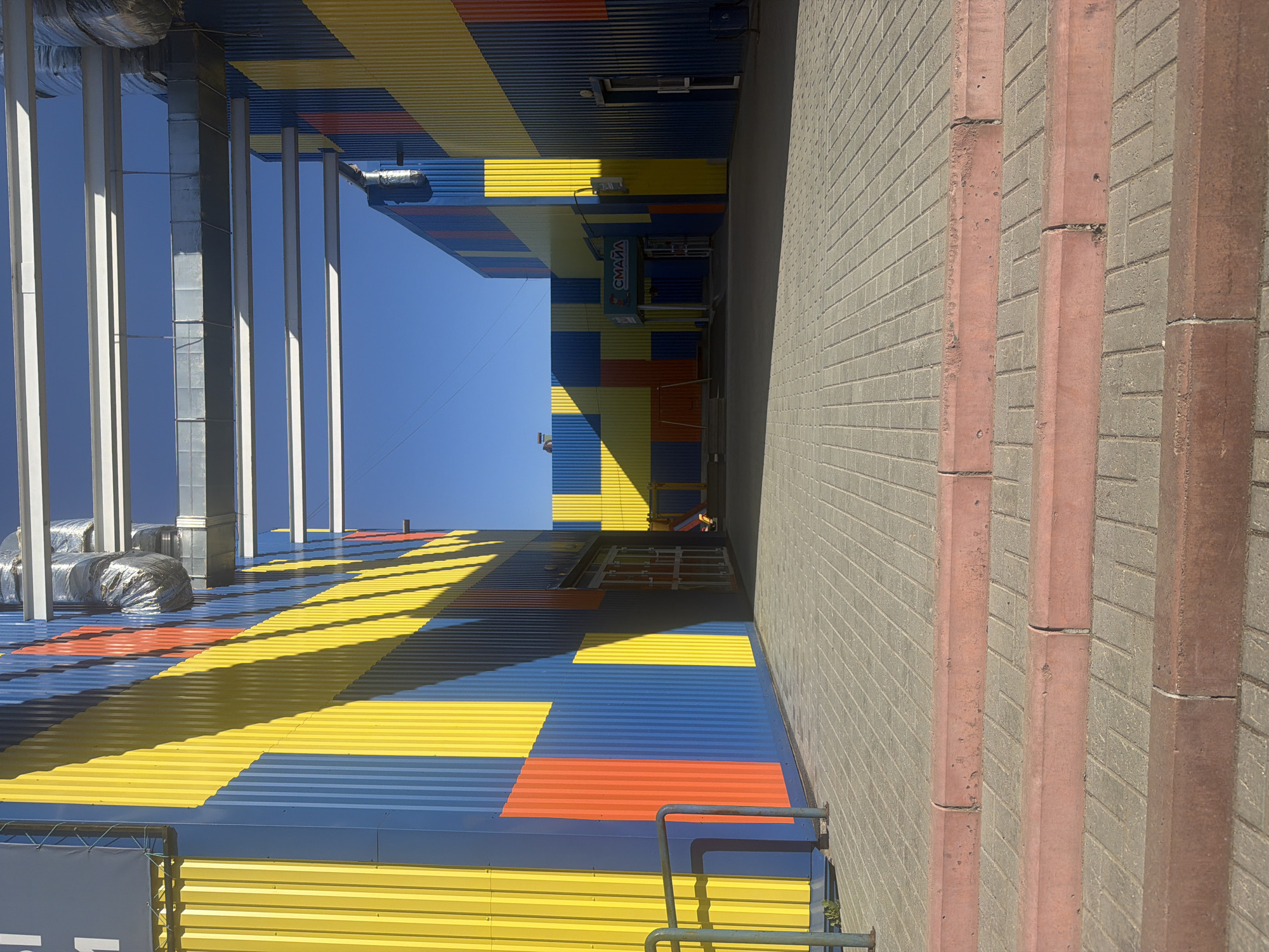 The image depicts the exterior of a building in Chaikovsky, Russia. The building has a colorful facade featuring blue, yellow, and orange horizontal stripes. The sign displays the word "Смайл" (Smile). There is a gray brick walkway alongside the building with vertical red brick accents. The sky is clear and blue. The image appears to be taken from ground level.