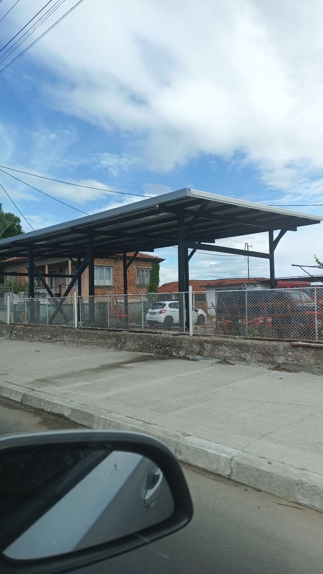The image shows a street scene in Milevo, Bulgaria, with a view of a car parked behind a chain-link fence, under a large metal canopy structure. Behind the fence, there is a low brick building with windows. A side mirror of a vehicle is visible in the foreground. The sky is partly cloudy. The ground is concrete. There are power lines overhead.