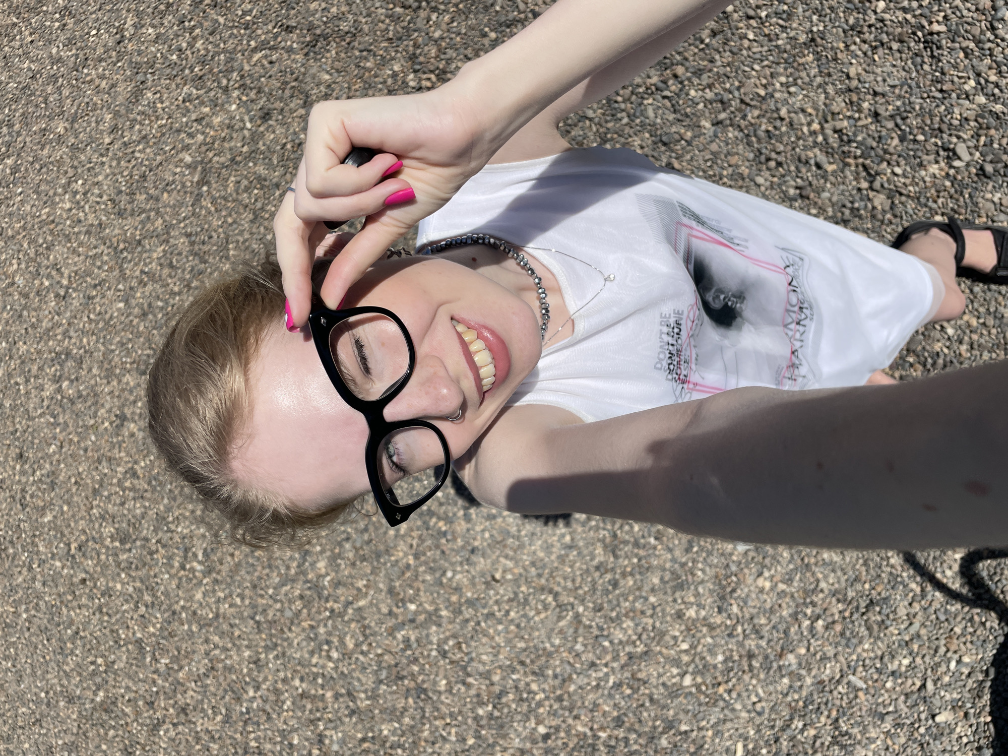 A pale-skinned person with blonde hair and wearing black-rimmed glasses, a silver necklace, and a white t-shirt with a print is lying on a pebbled surface. The person's right hand is adjusting the glasses, and the fingernails are painted bright pink. The person is smiling, revealing their teeth. The person's feet wearing sandals can be seen on the right side of the frame. The surface appears to be asphalt or gravel, suggesting an outdoor location. The scene is brightly lit, implying a sunny day. There are no indications to determine the location in Izhevsk, Russia.