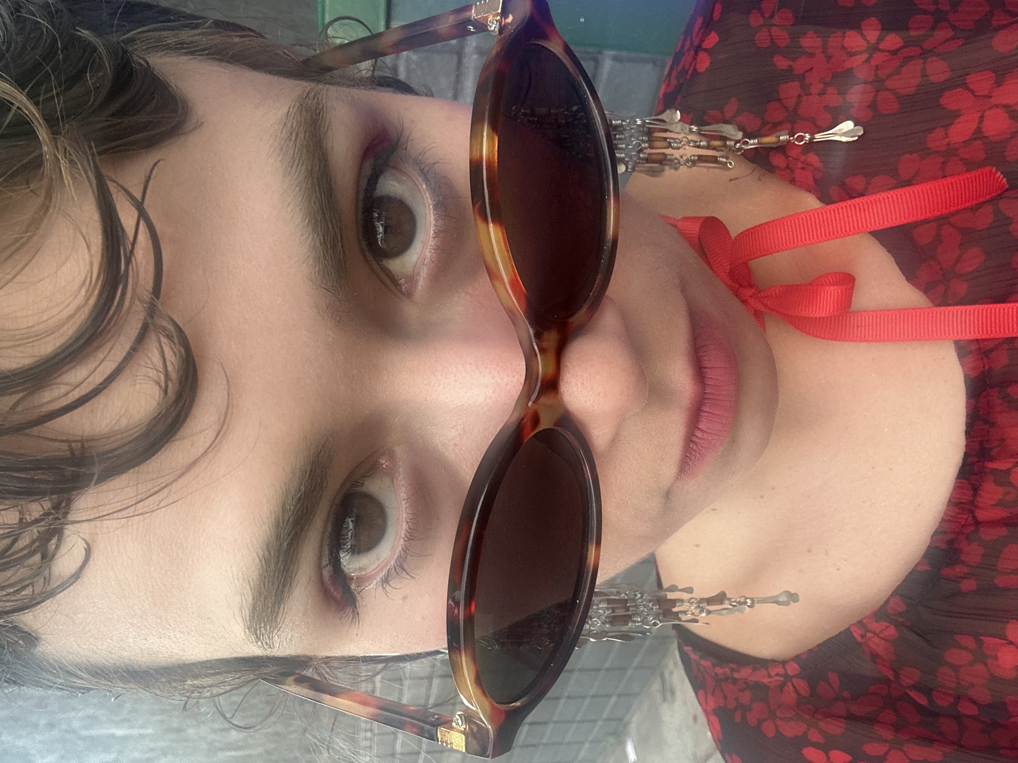 A close-up, slightly rotated shot features a person wearing sunglasses and a red floral-patterned top. The sunglasses are tortoiseshell-patterned with dark lenses. A red ribbon is tied around the person's neck. The person's eyes are visible above and below the sunglasses. Attached to the sunglasses are dangling silver-colored accessories. The person's hair is styled in waves. The scene does not give sufficient clues to identify a specific location in Los Angeles.