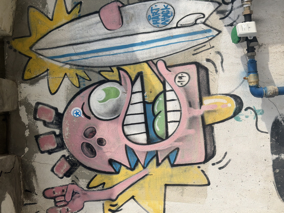 The image displays colorful graffiti on a textured, light-colored concrete or stucco wall in Tel-Aviv, Israel. The precise location within the city cannot be determined from the visual evidence.
The central artwork features a cartoonish, pink-skinned character with a spiky head and a wide-open mouth revealing numerous white teeth and a green tongue. On the left side of its head are three dark circular holes. Its right side displays a single large, bulging eye with a green iris and black pupil. A blue circular sticker, containing a white star and magnifying glass icon, is affixed to the character's forehead.
The character's left arm, also pink, is raised in a "rock on" hand gesture. Its right arm holds a white surfboard vertically. The surfboard has two blue vertical stripes and a circular logo with "HART ATTACK" in blue text. Yellow, jagged, starburst-like shapes emanate from behind the character and the surfboard.
Below the graffiti on the lower right, a blue PVC pipe system is affixed to the wall, incorporating a brass valve and a white utility device with a green cap. The wall itself shows surface texture, minor cracks, and some discoloration. Faint blue lines of previous graffiti are visible beneath the primary artwork.